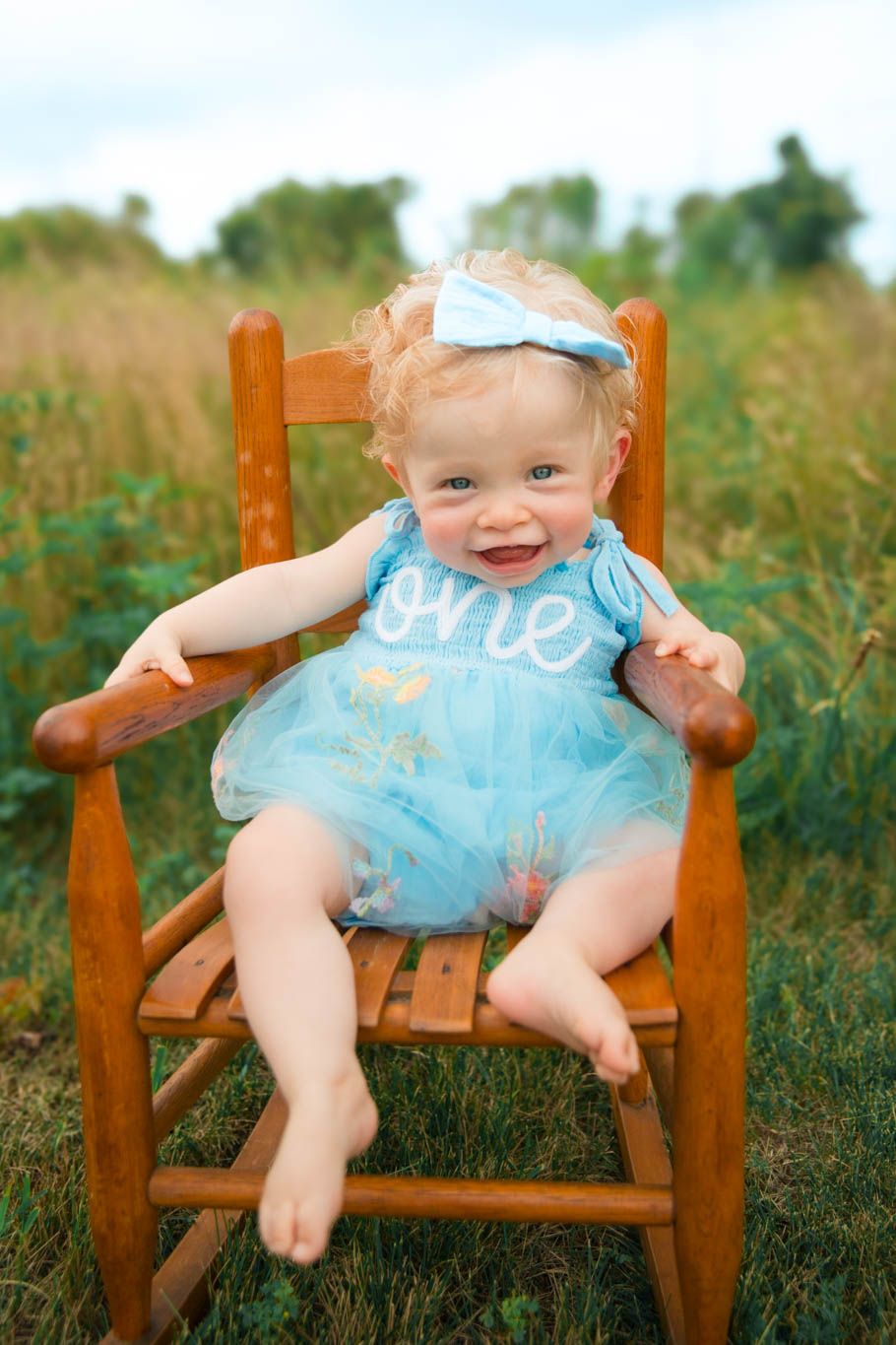 Baby wearing blue tulle dress sits in wooden rocking chair outdoors in field of tall grass.
