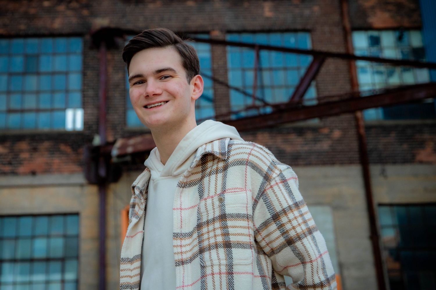 Person wearing plaid hoodie smiles in front of industrial brick building.