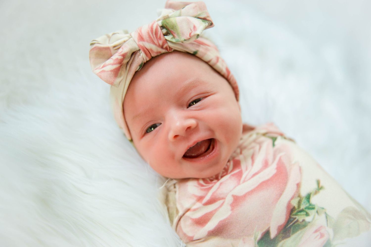 Baby wearing floral outfit and matching headband smiles on white background.