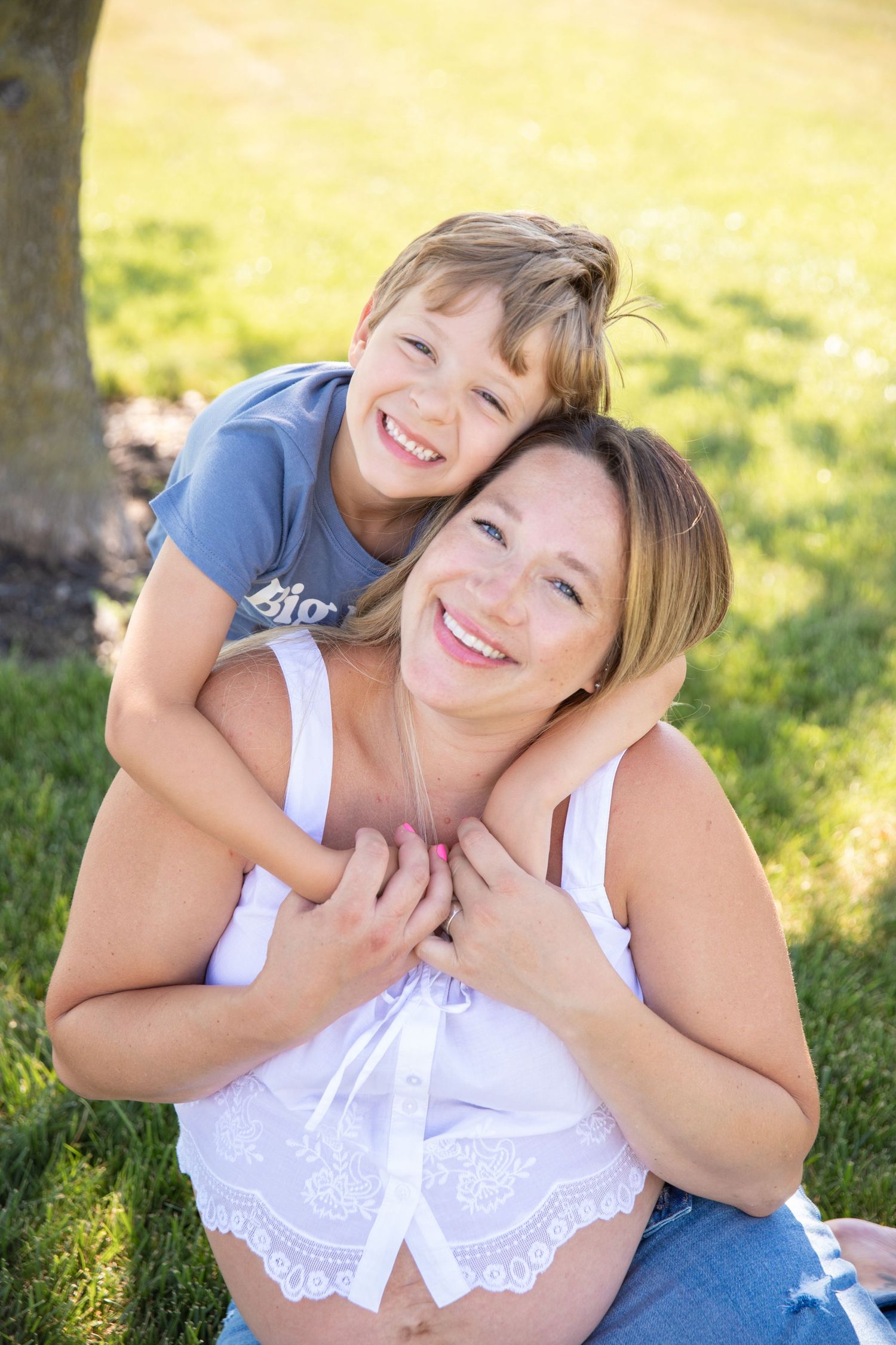 Happy pregnant mother and young son hugging outdoors in green park, smiling together on sunny day.