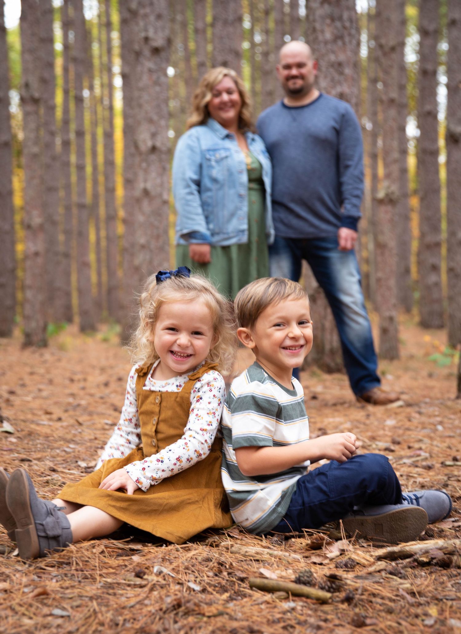 Children sit on fallen leaves while parents stand behind in pine forest setting.
