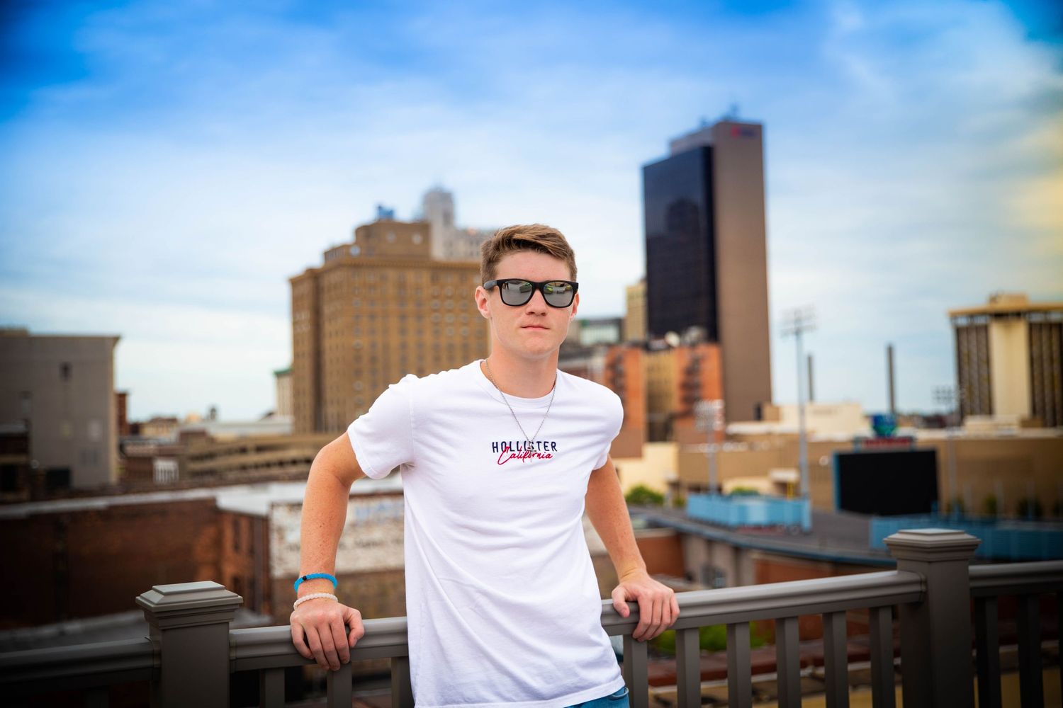 Person in white t-shirt poses on urban rooftop with city skyline backdrop.