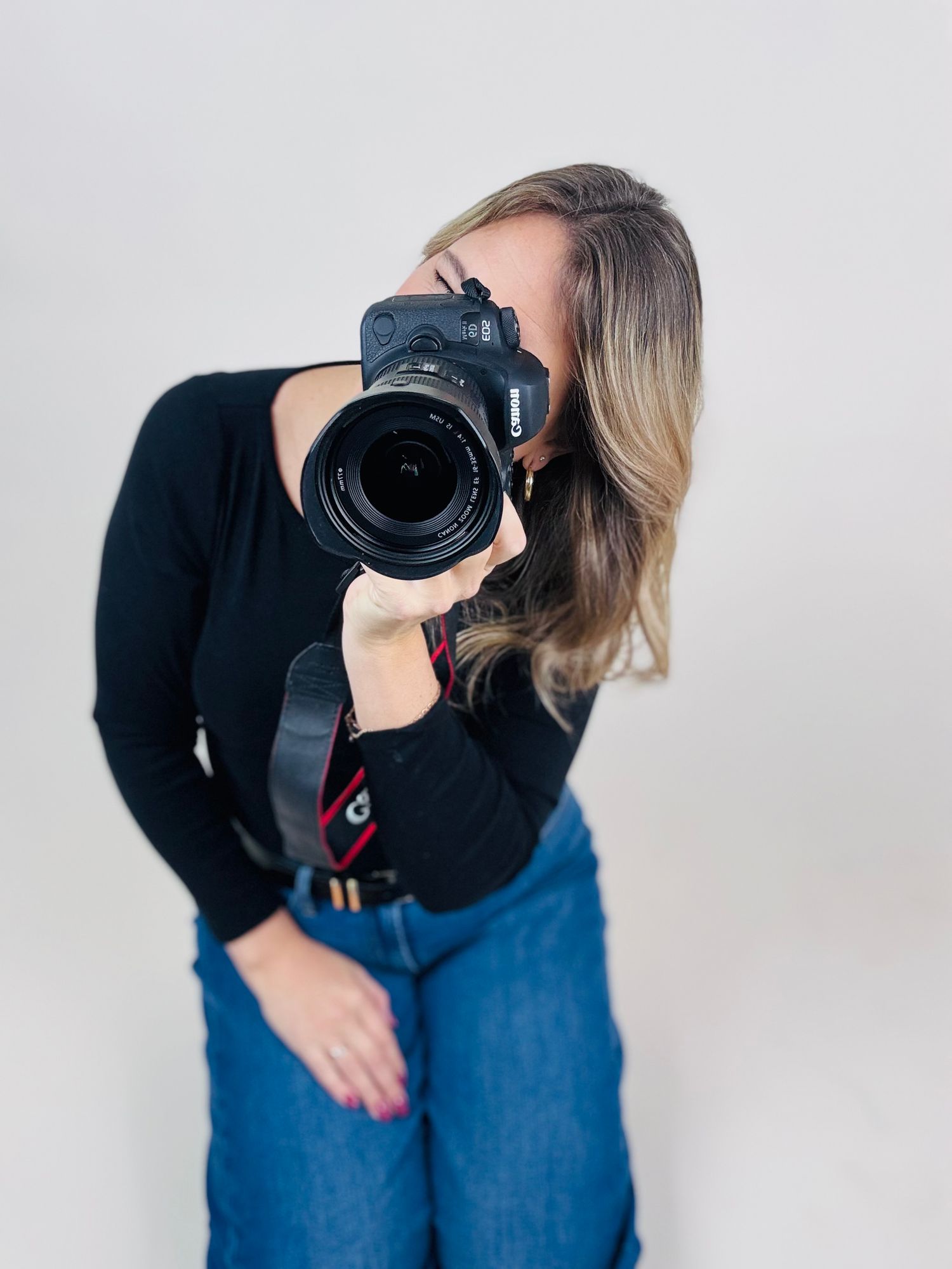 Professional photographer wearing black sweater and jeans demonstrates four different camera holding positions against white wall.