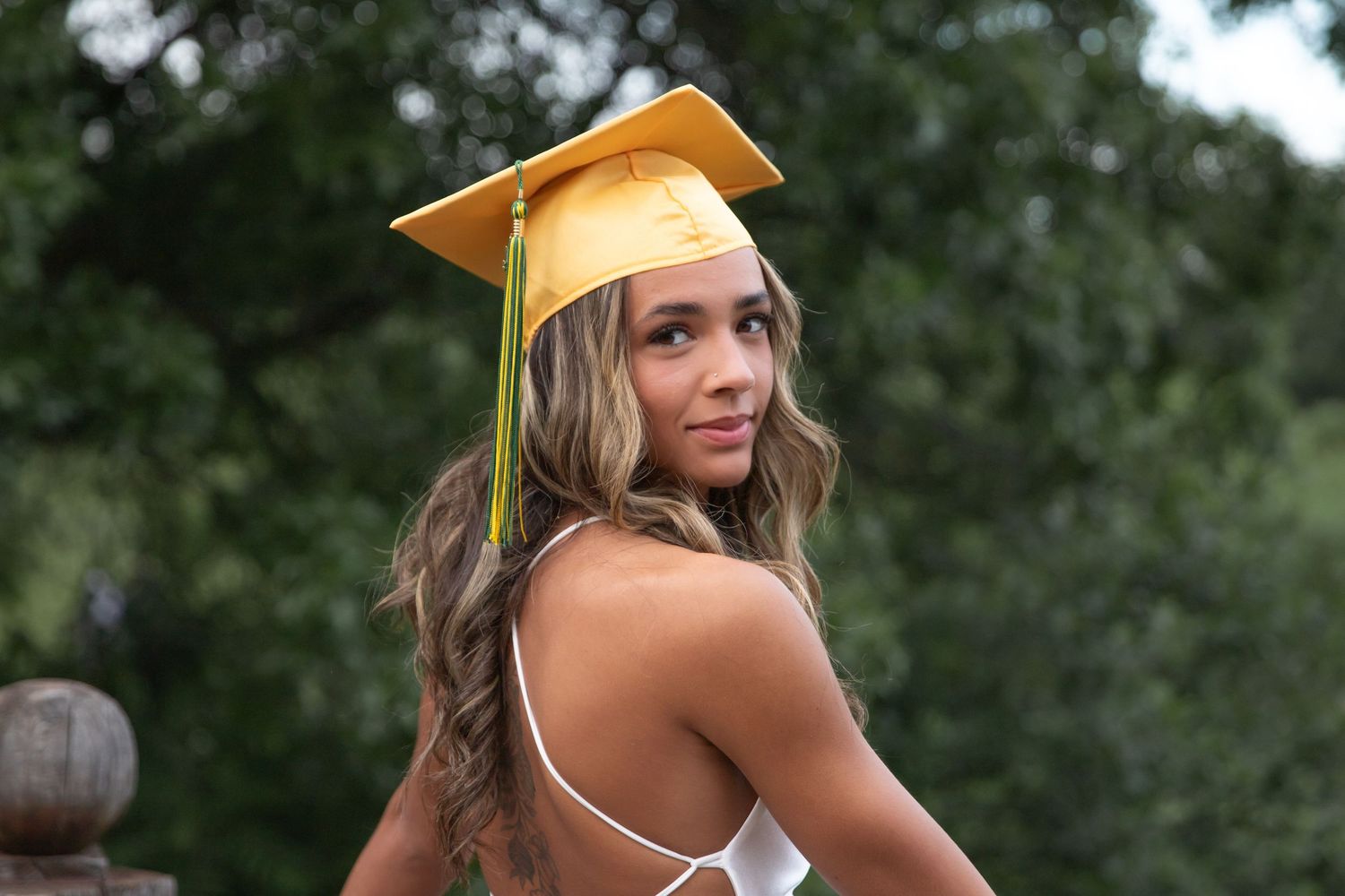 Graduate in yellow cap and white dress poses outdoors during graduation photoshoot.