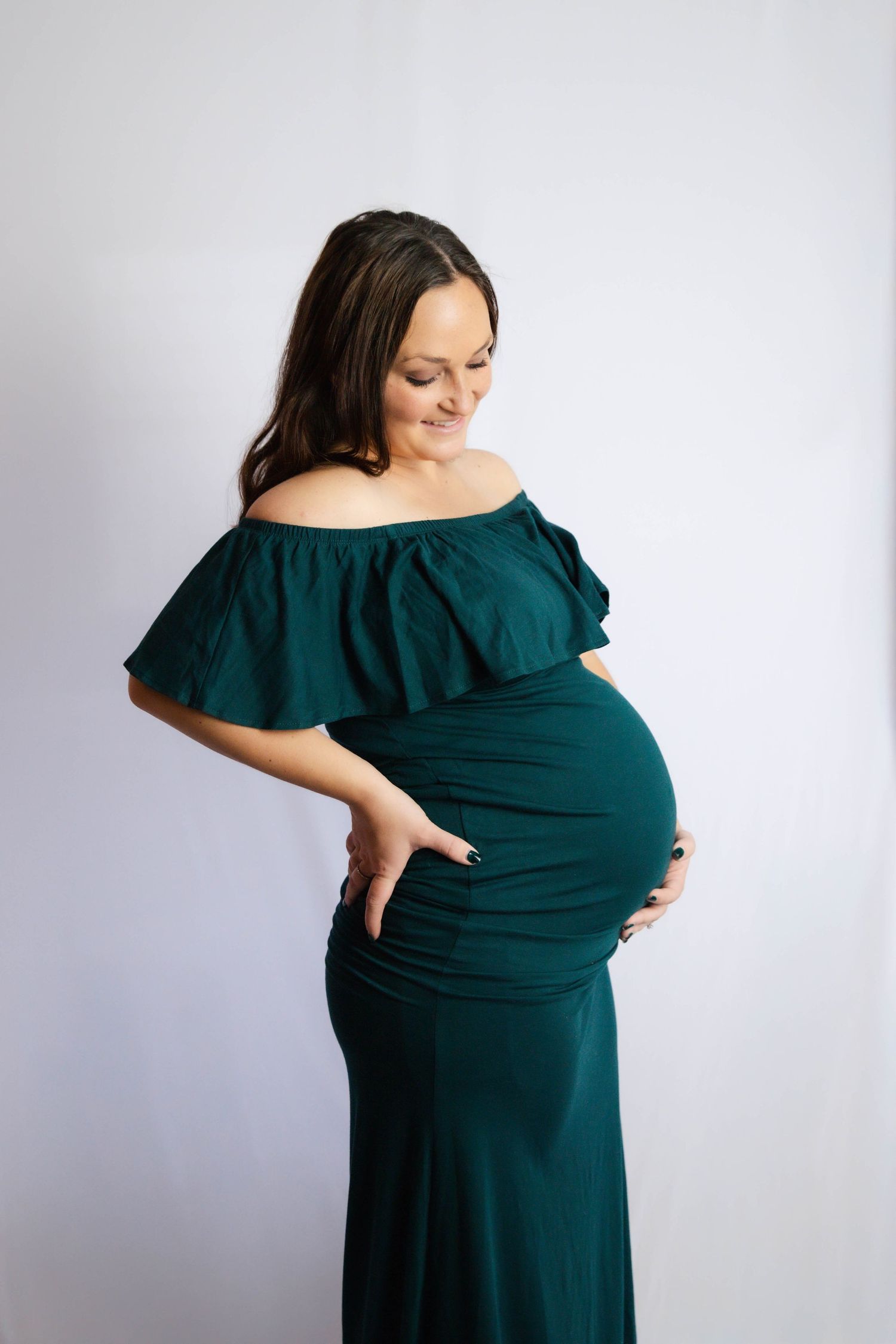 Maternity photo series showing woman in emerald dress cradling baby bump against white background.
