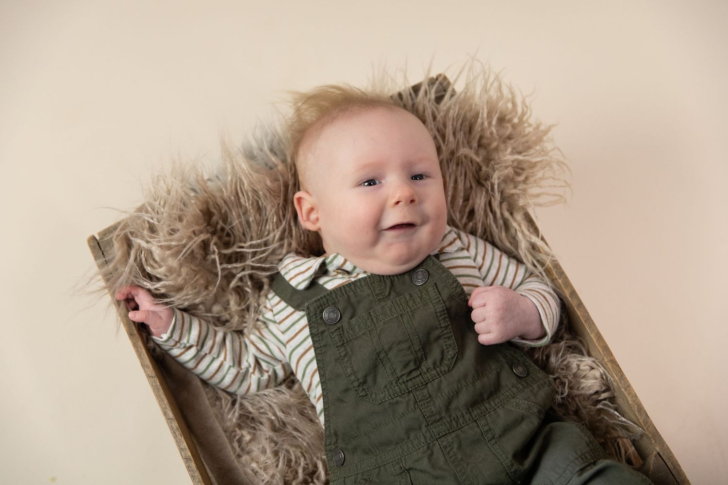 Smiling baby boy in green overalls and striped shirt lying on fluffy fur in a wooden box.