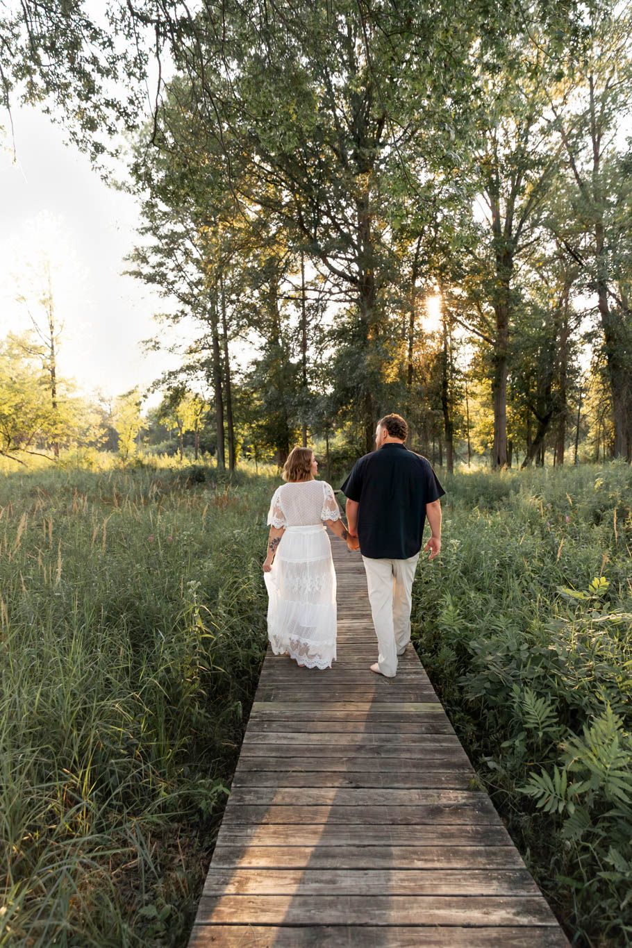 Couple walking together on wooden boardwalk through natural prairie at sunset.