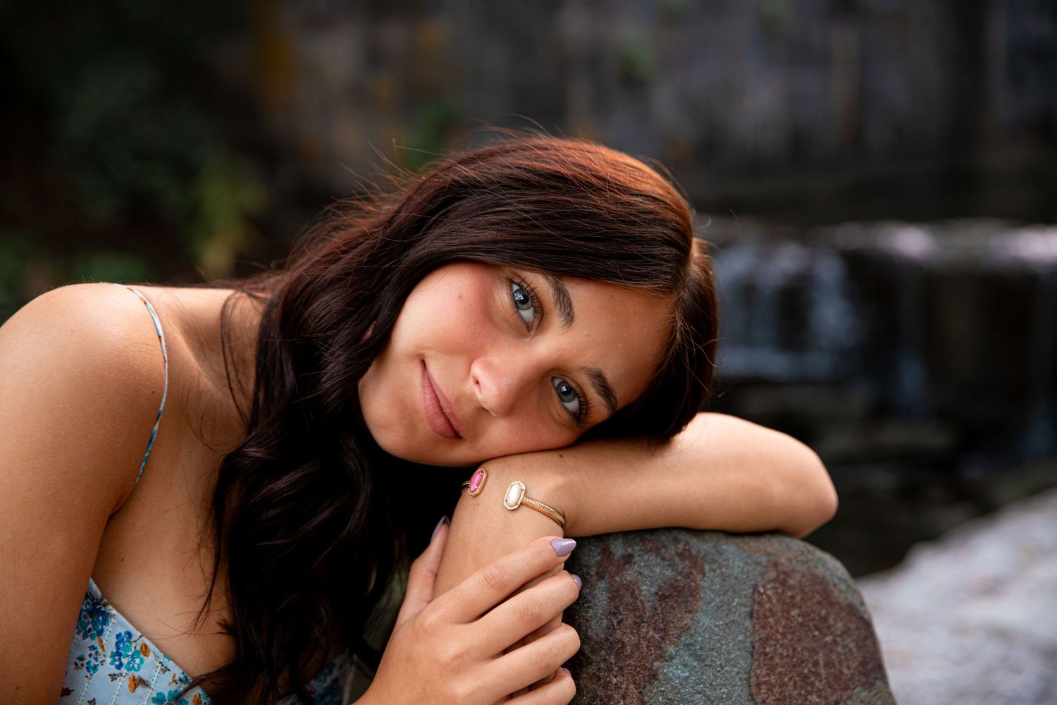 Young woman with dark hair resting her chin on hands, wearing a floral top and gold ring outdoors.
