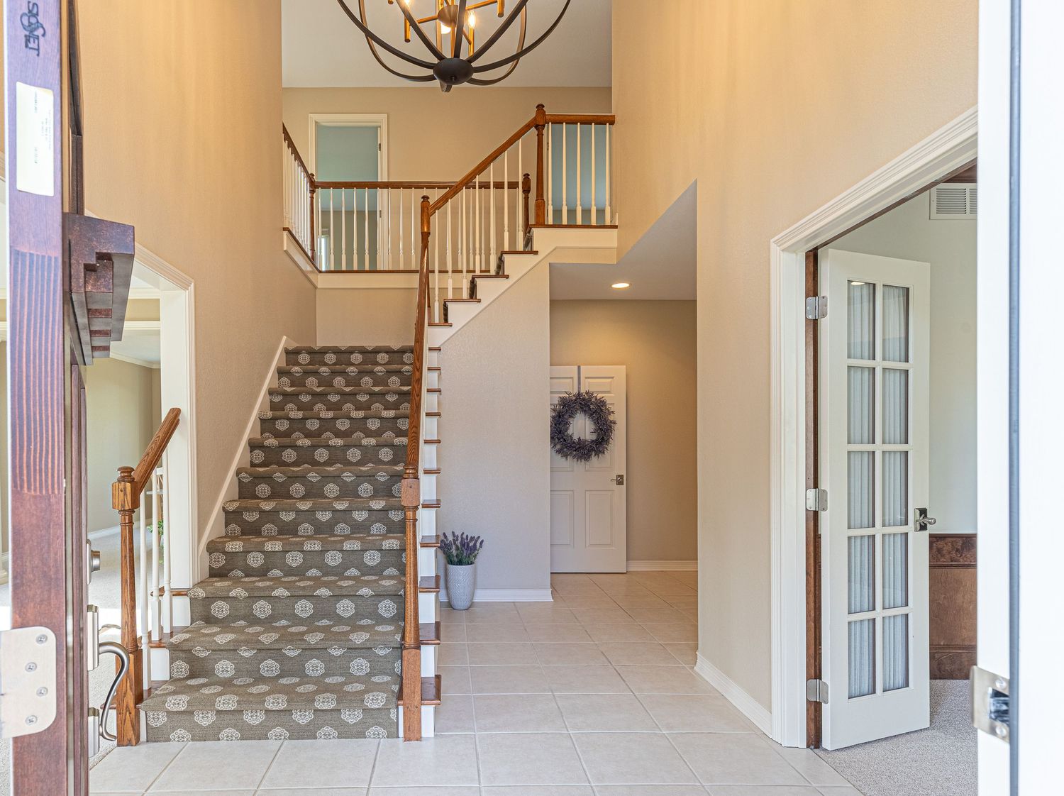 Bright two-story foyer with patterned staircase carpet, chandelier, white walls, and French door leading to adjacent room.