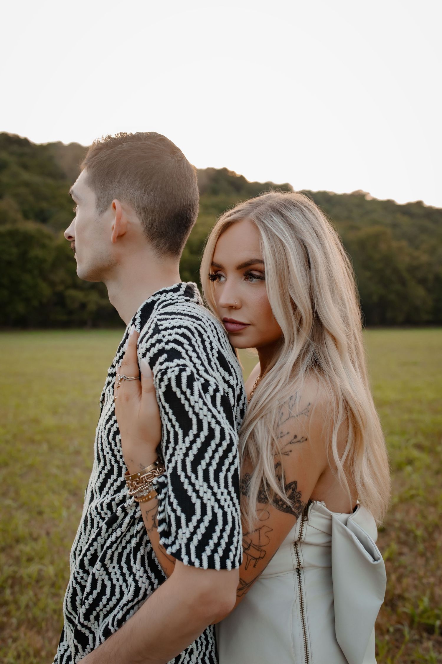 A blonde woman with tattoos embraces a man in a black and white patterned shirt in a green field at dusk.