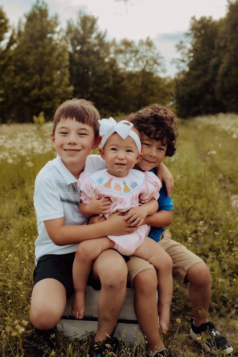 Two brothers smile while holding their baby sister in a sunny meadow.