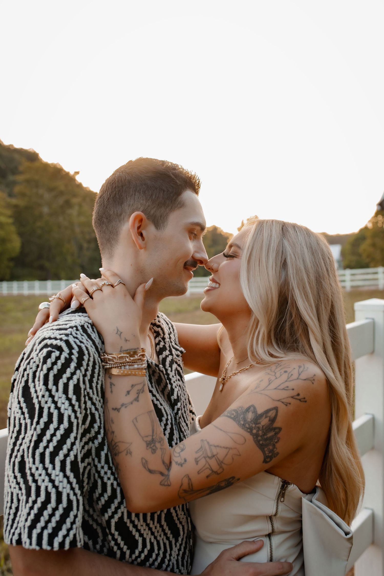 Tattooed couple sharing a romantic moment outdoors at golden hour, smiling and embracing near a white fence.