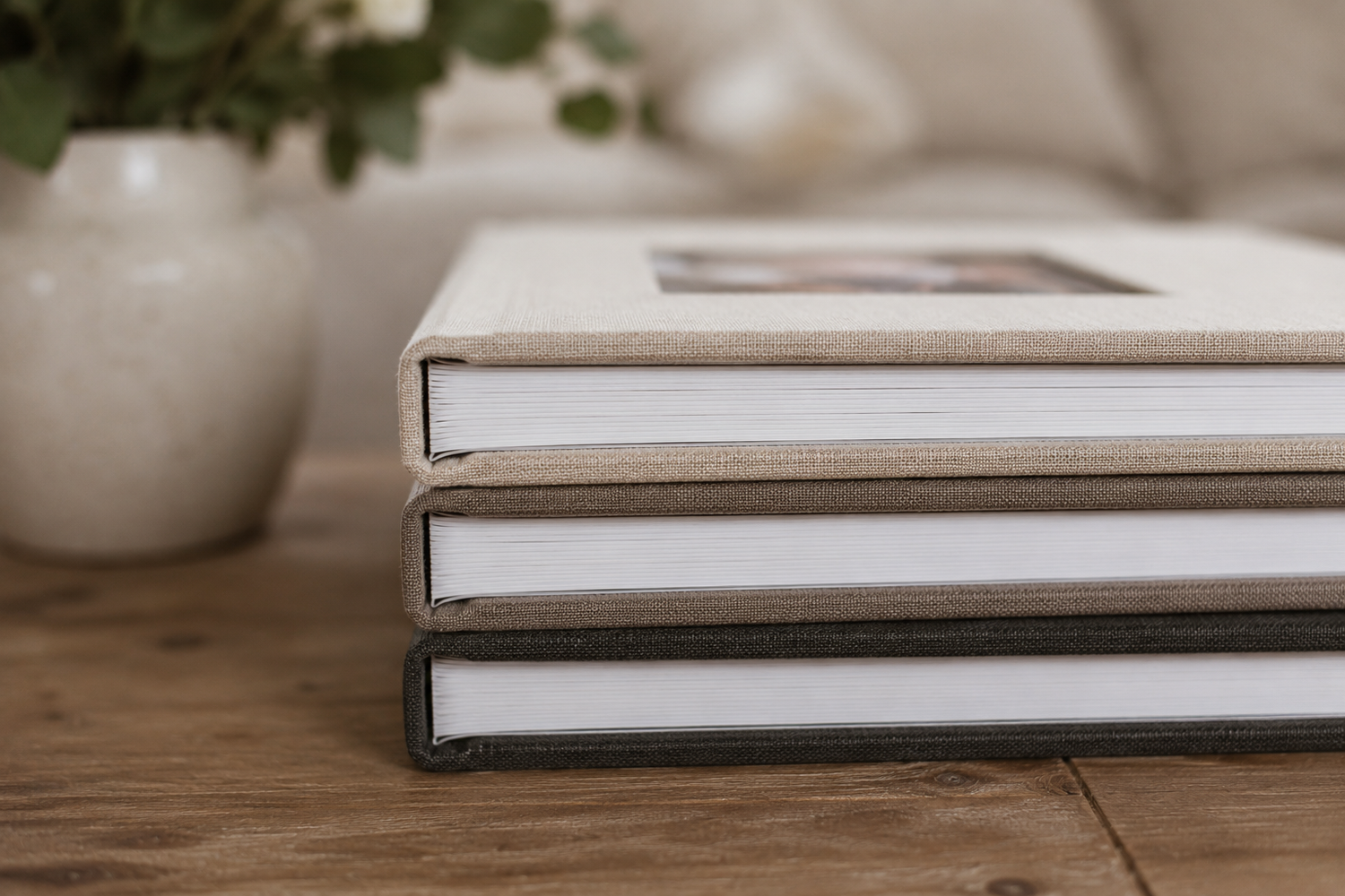 Stack of three elegant hardcover photo albums on a wooden table with a white vase in the background.