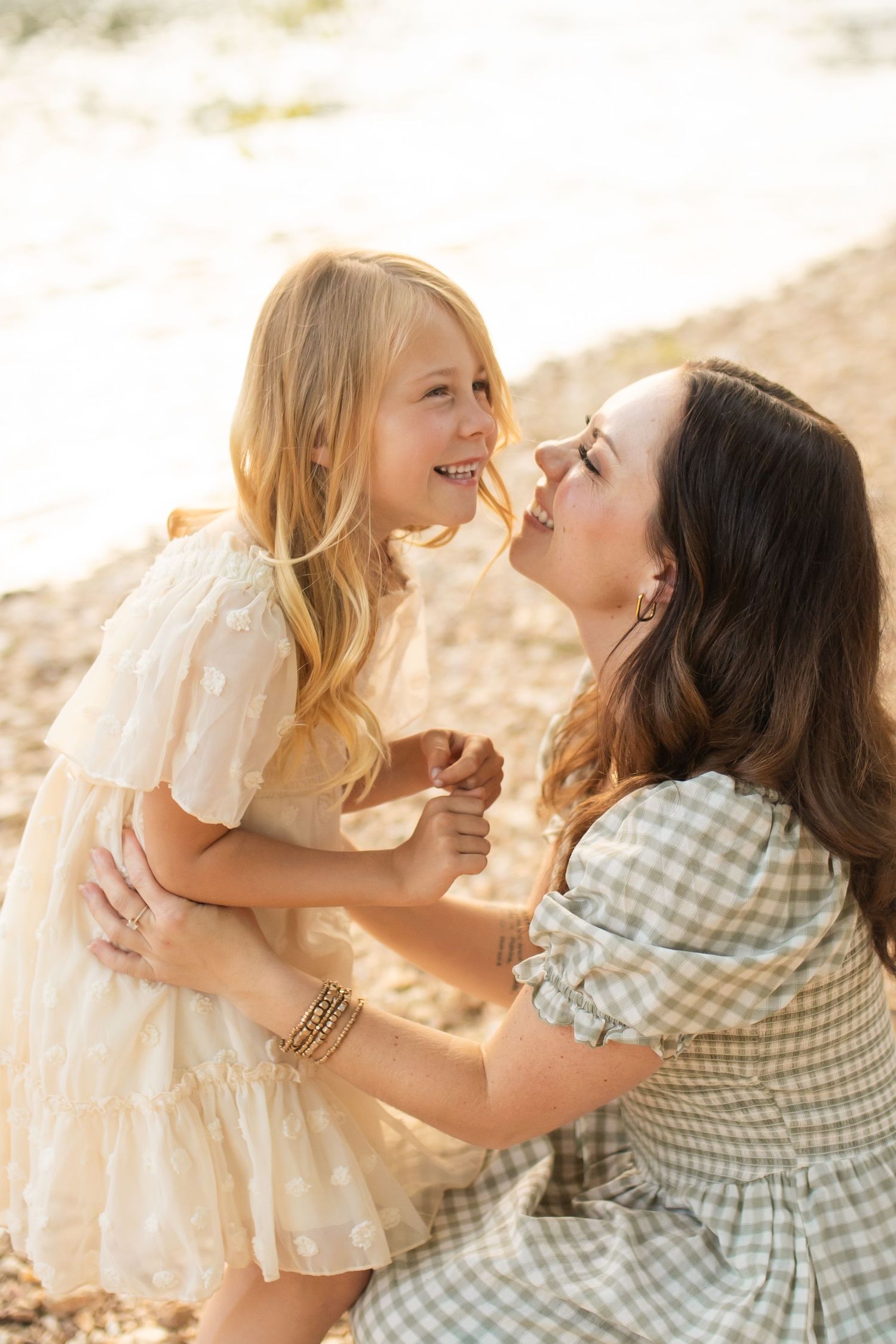 A mother and young blonde girl in a cream dress share a joyful nose-to-nose moment on a sunny beach.