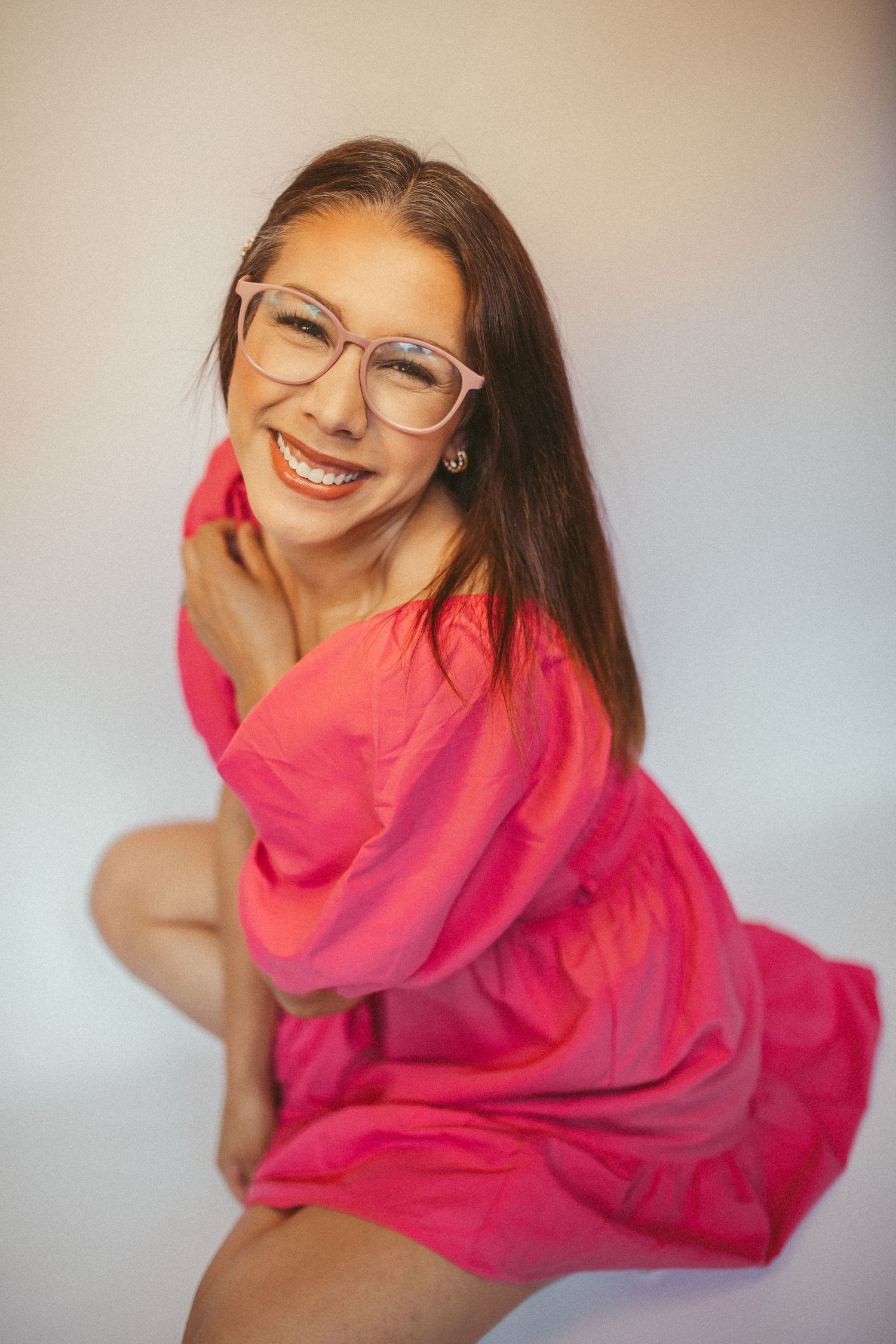 Smiling woman in pink dress and glasses posing playfully against a white background.