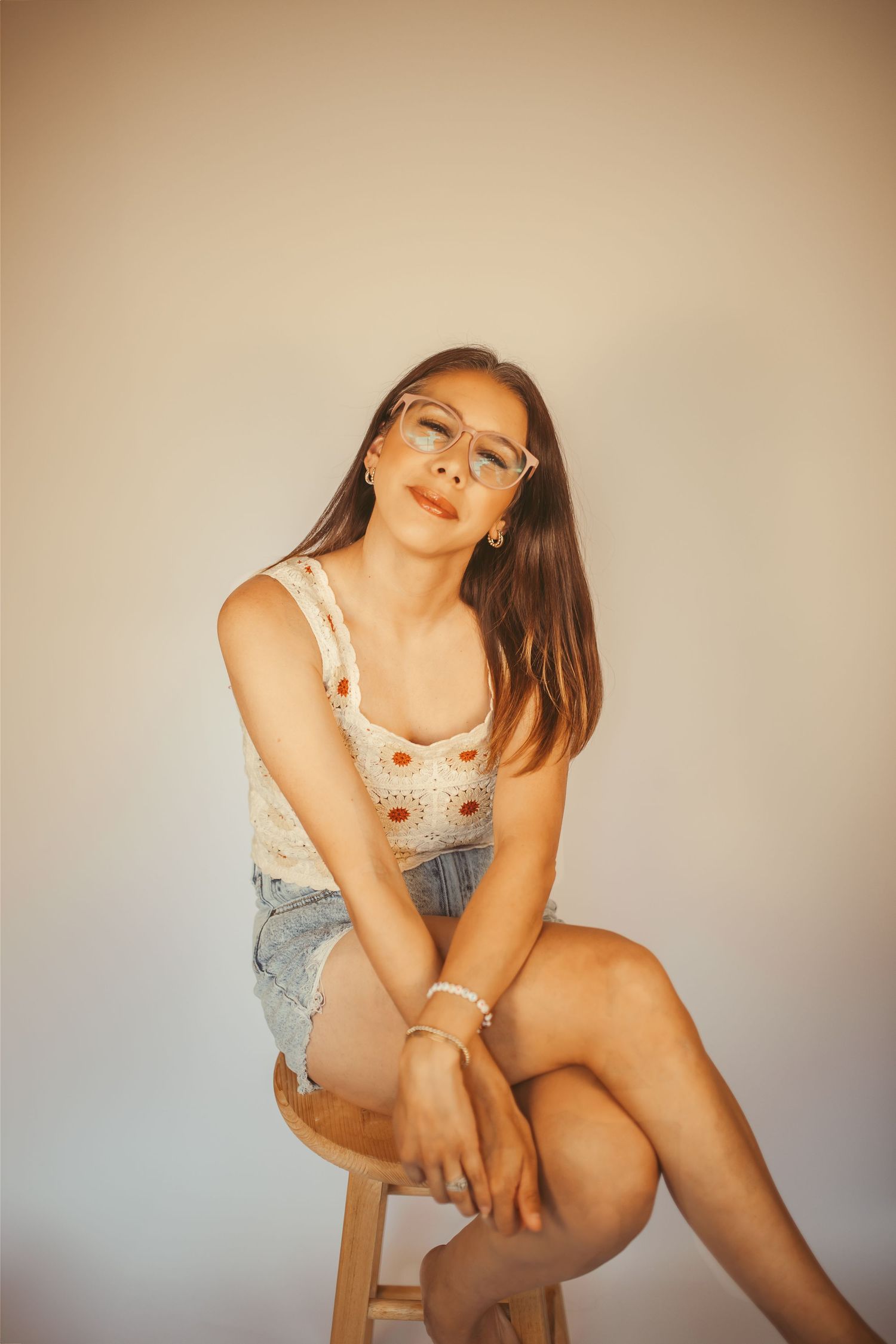 Young woman in denim shorts and floral top sitting on stool, wearing clear glasses, posing against neutral background in Ozark, Mo.
