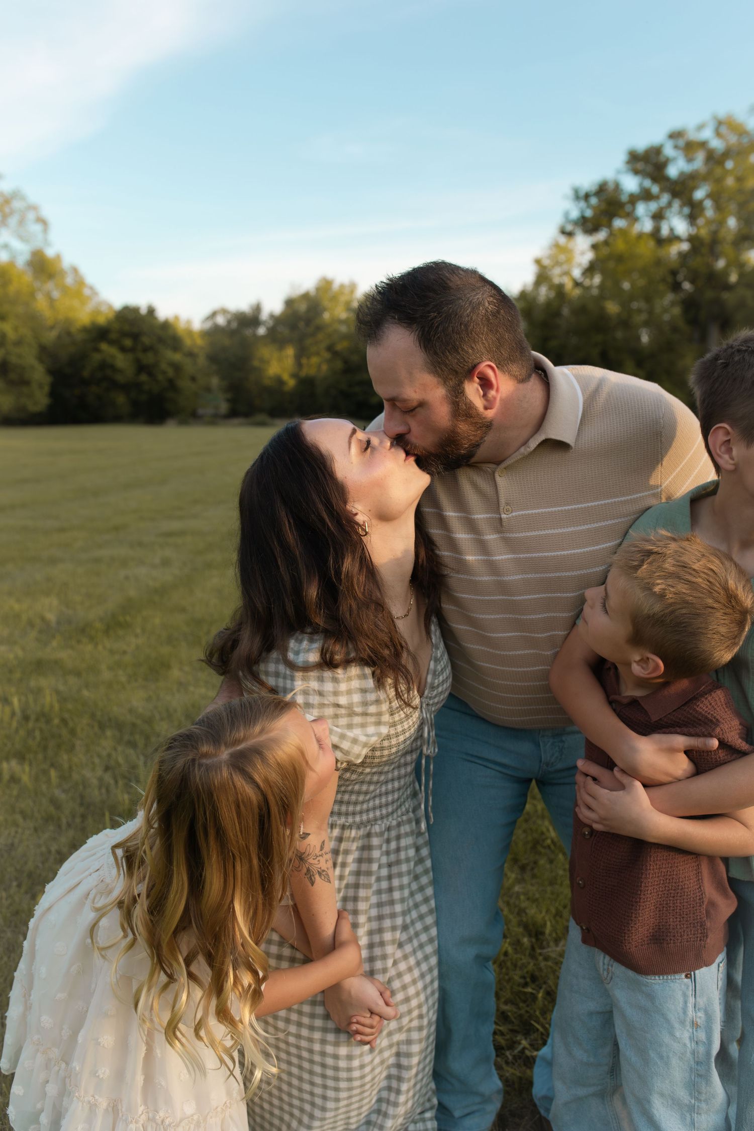 A couple shares a kiss in a sunlit field while holding their two young children during a family portrait session.
