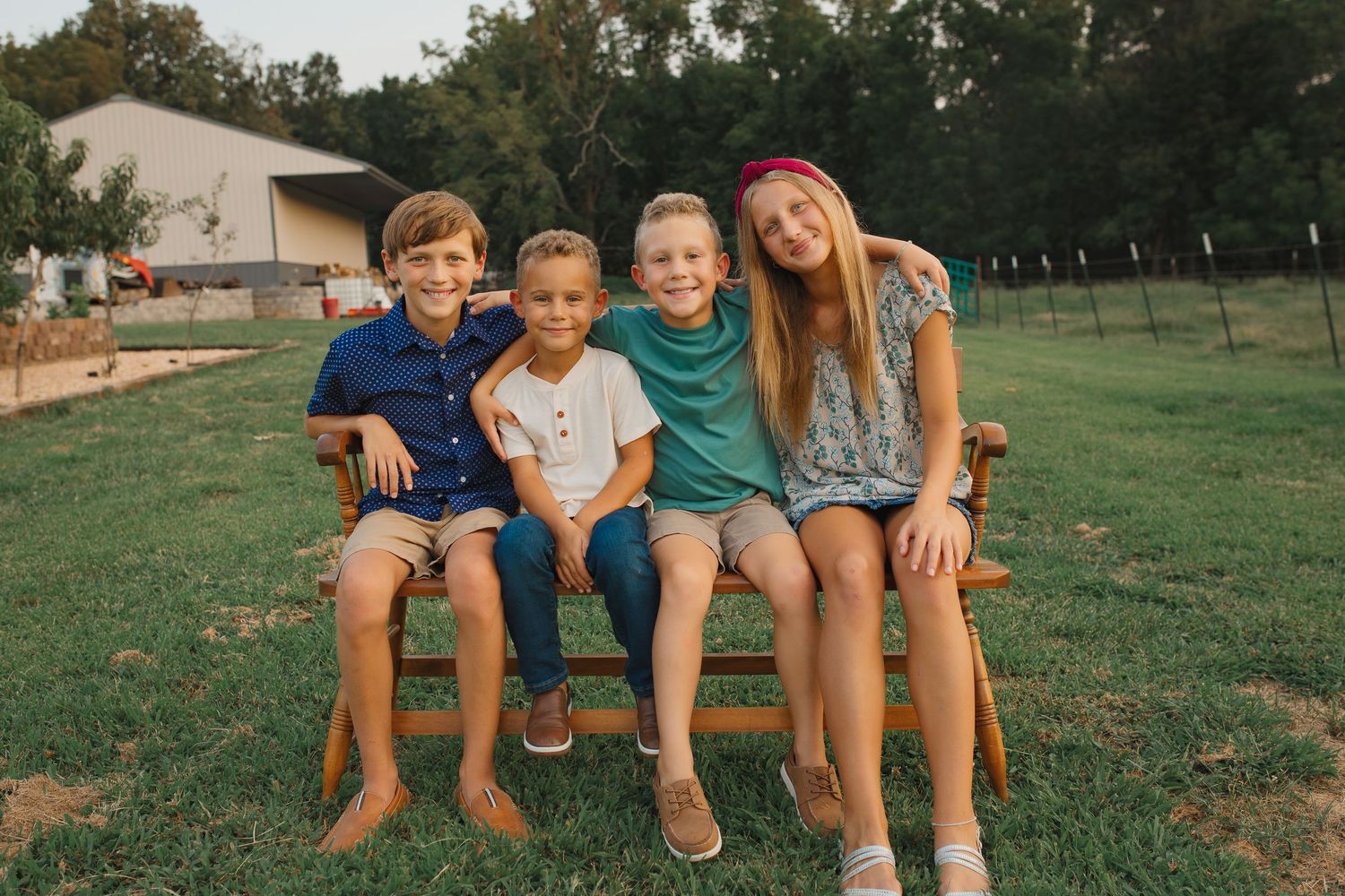 Four children sitting together on a wooden bench outdoors on a farm, smiling for a portrait photo.