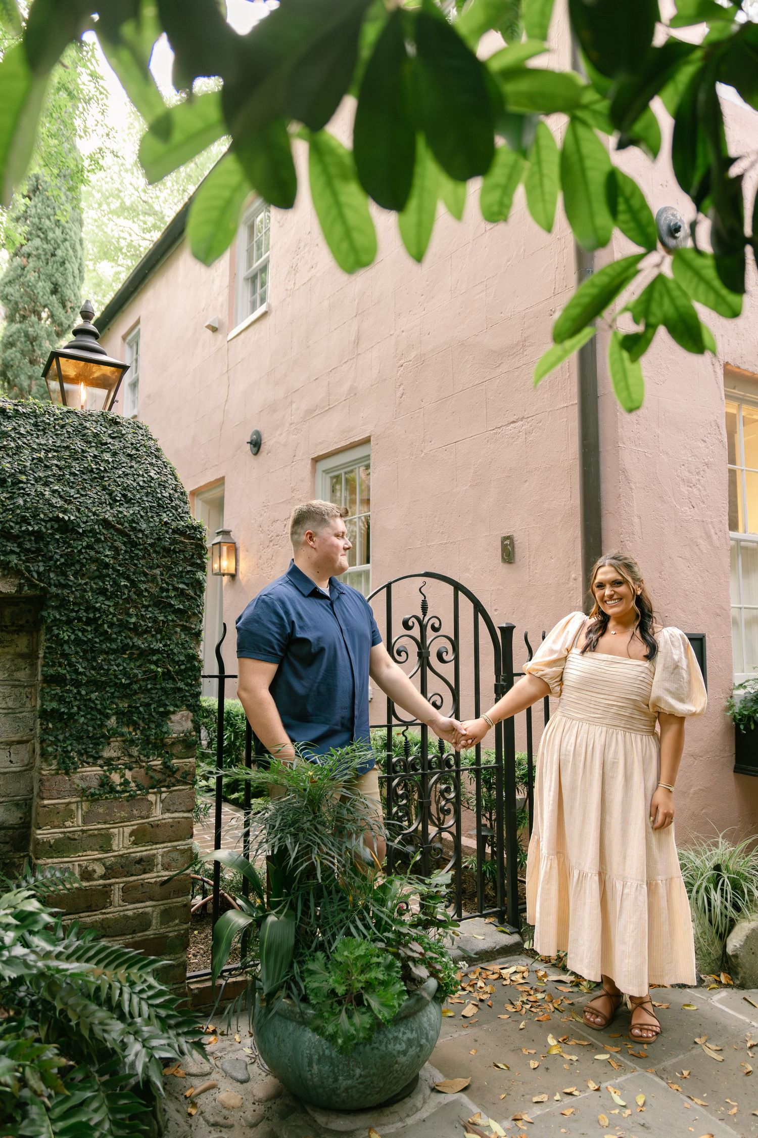 Couple holding hands smiling at each other by iron gate with lush greenery and pink building backdrop.