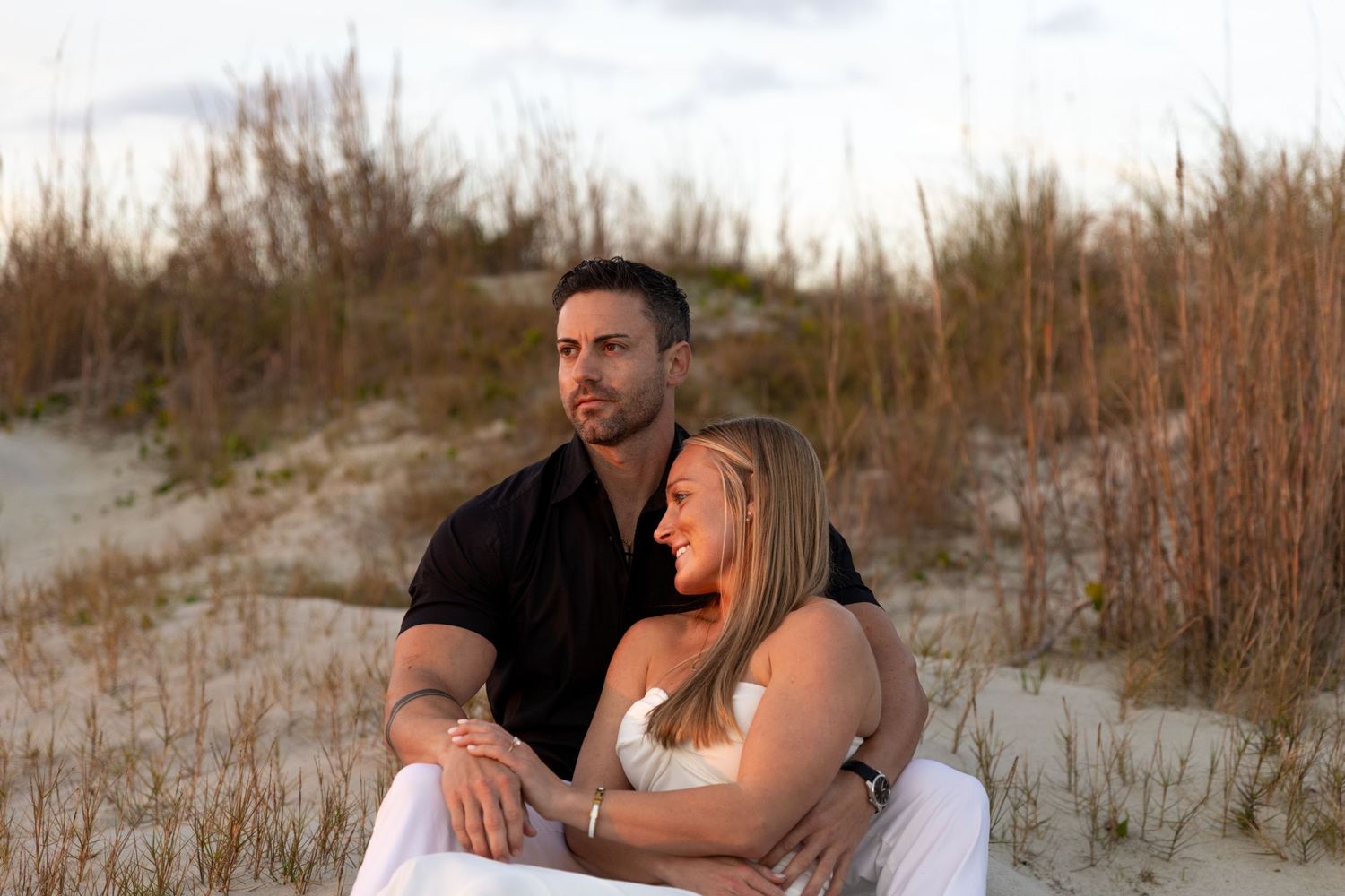 Couple sitting together on sandy beach dunes surrounded by tall grass during golden hour sunset.