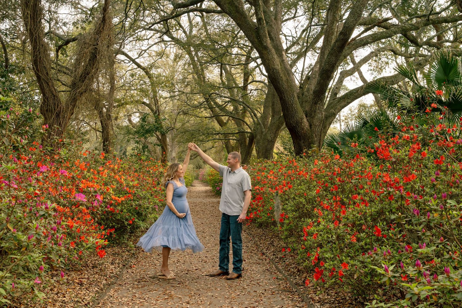 Couple dancing among vibrant red and pink flowers on a garden path surrounded by large oak trees.