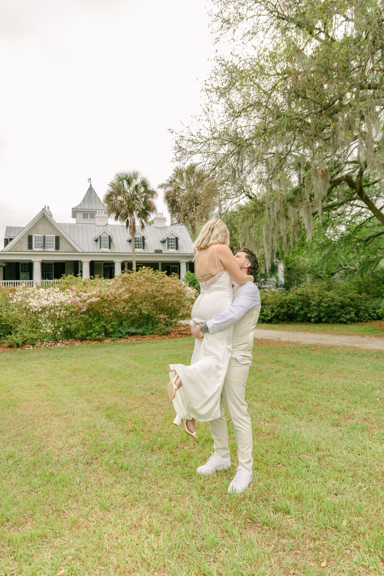 Groom lifts bride in white dress outdoors on green lawn before elegant Southern plantation home with Spanish moss trees.