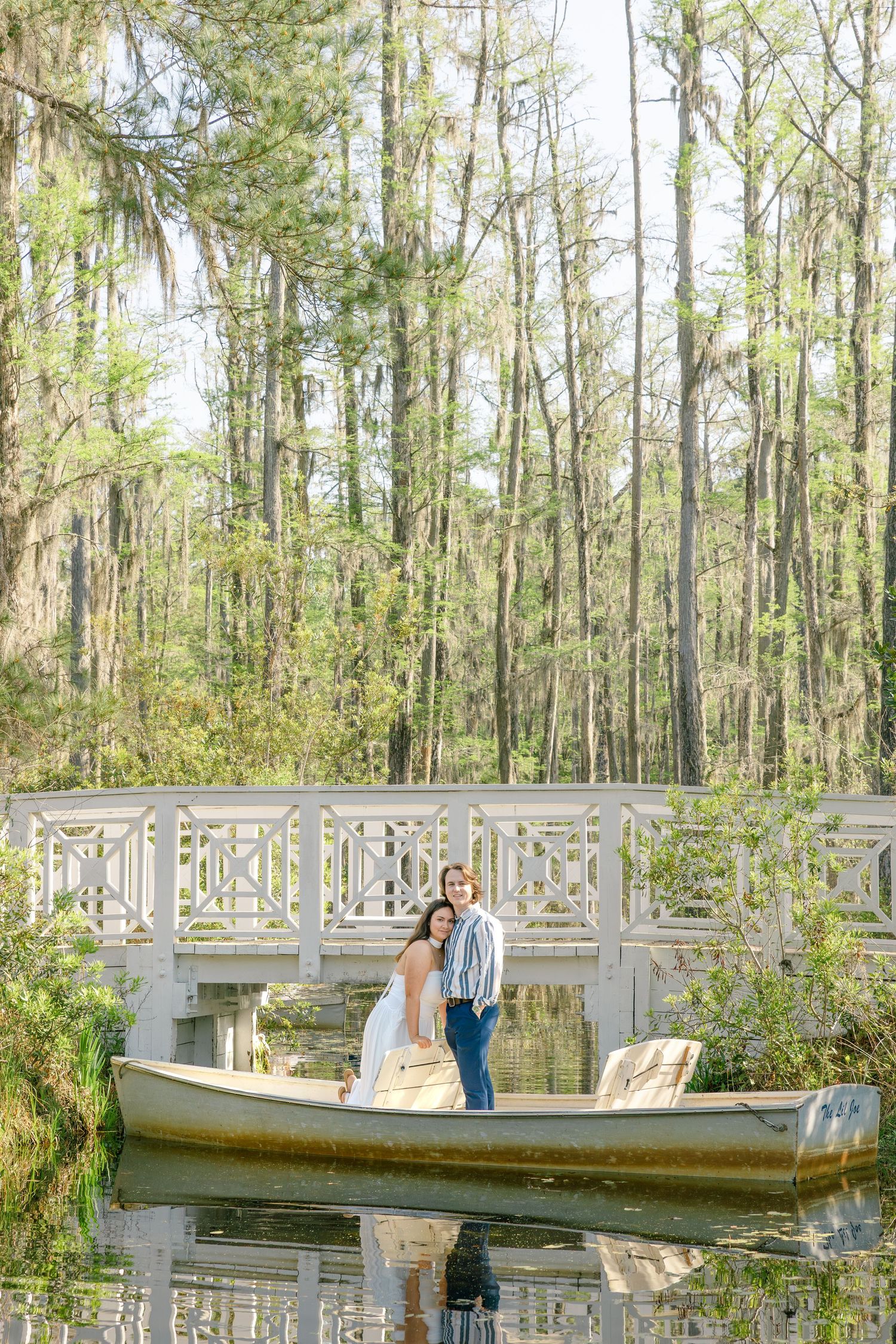 Couple in a canoe on a tranquil pond, posing near a white bridge surrounded by lush green trees.