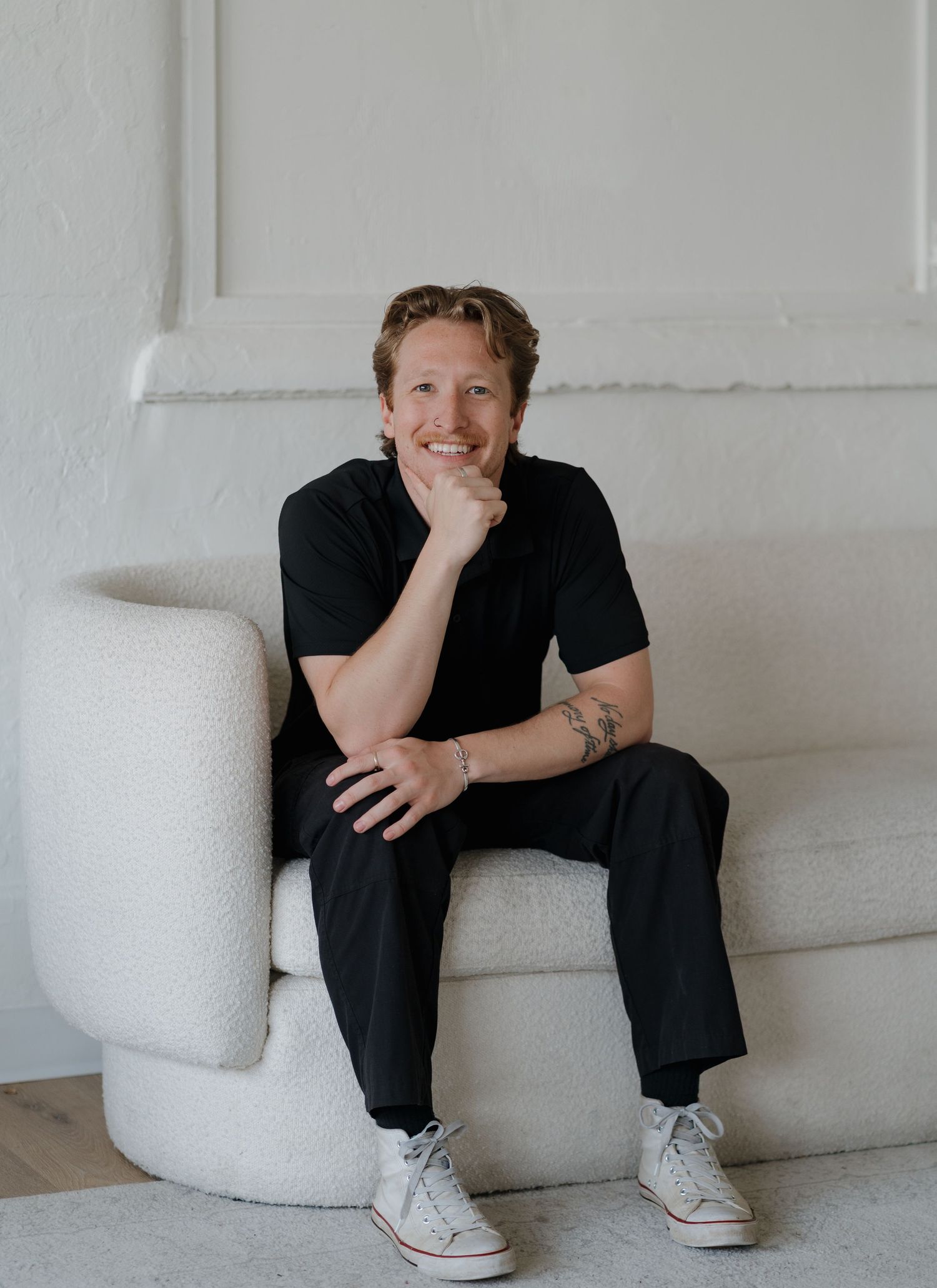 A person wearing black clothing and white sneakers rests casually on a white modern bench against a light colored wall.