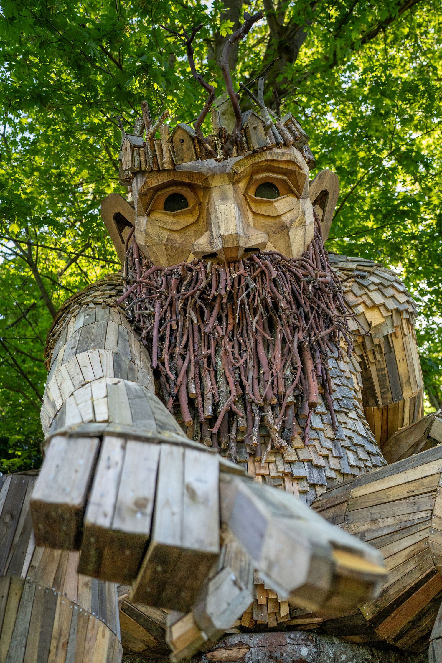 Large wooden sculpture of a face with rustic beard detail among green trees.