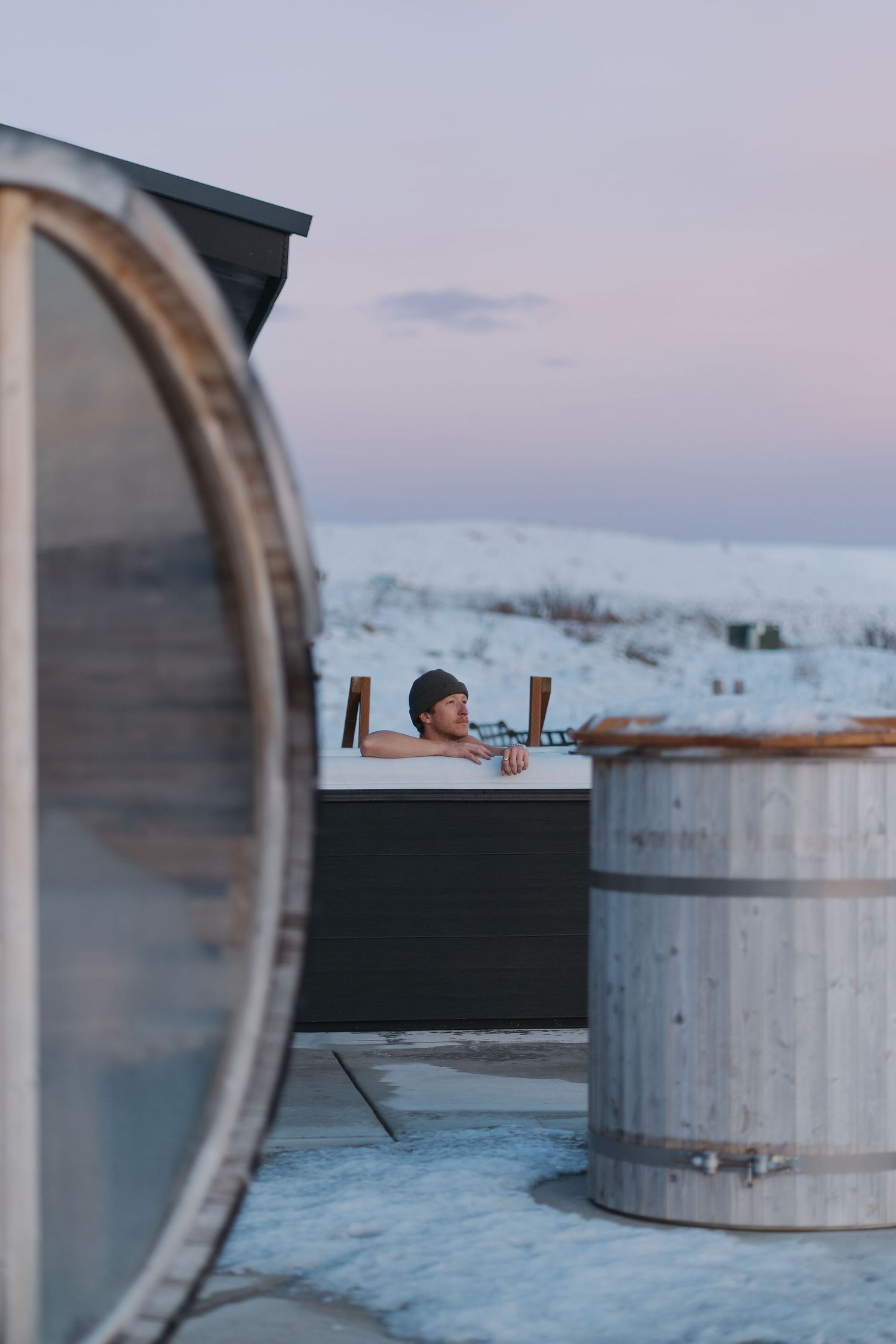 A person relaxes in an outdoor hot tub surrounded by snowy landscape during winter twilight.