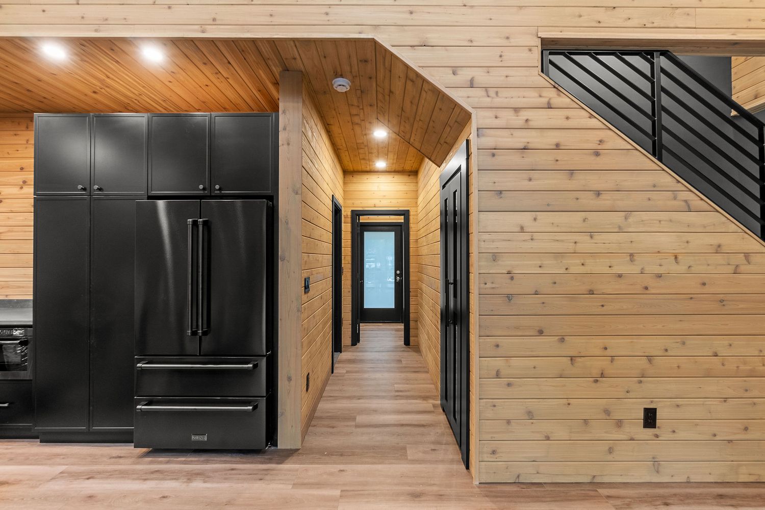 Modern kitchen hallway with black appliances and natural pine wood walls and flooring.