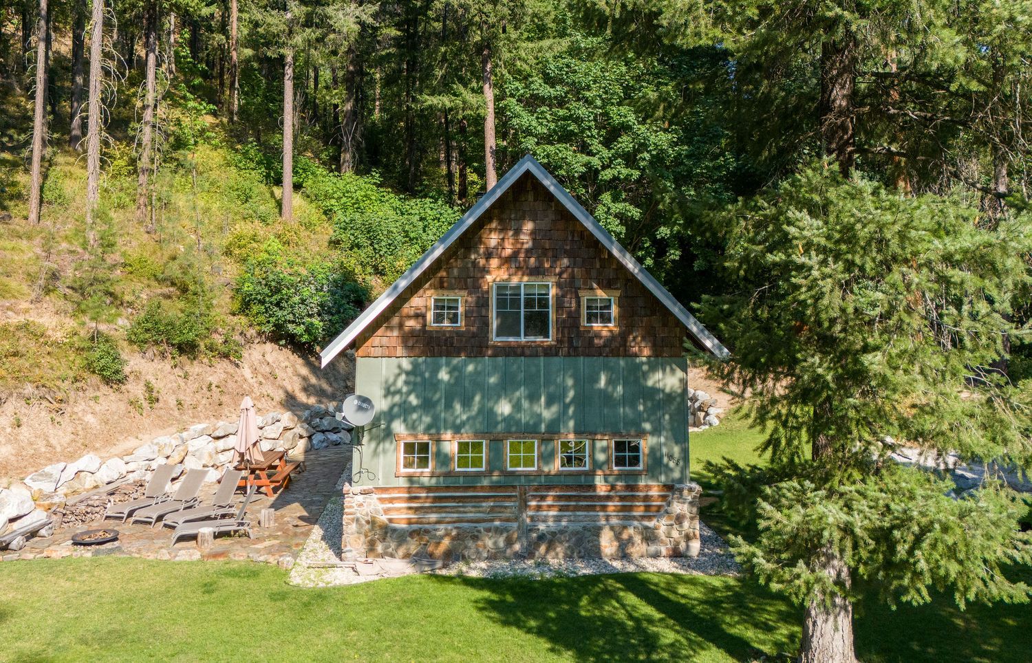 Aerial view of wooden cabin nestled in lush forest with green lawn and rock landscaping.