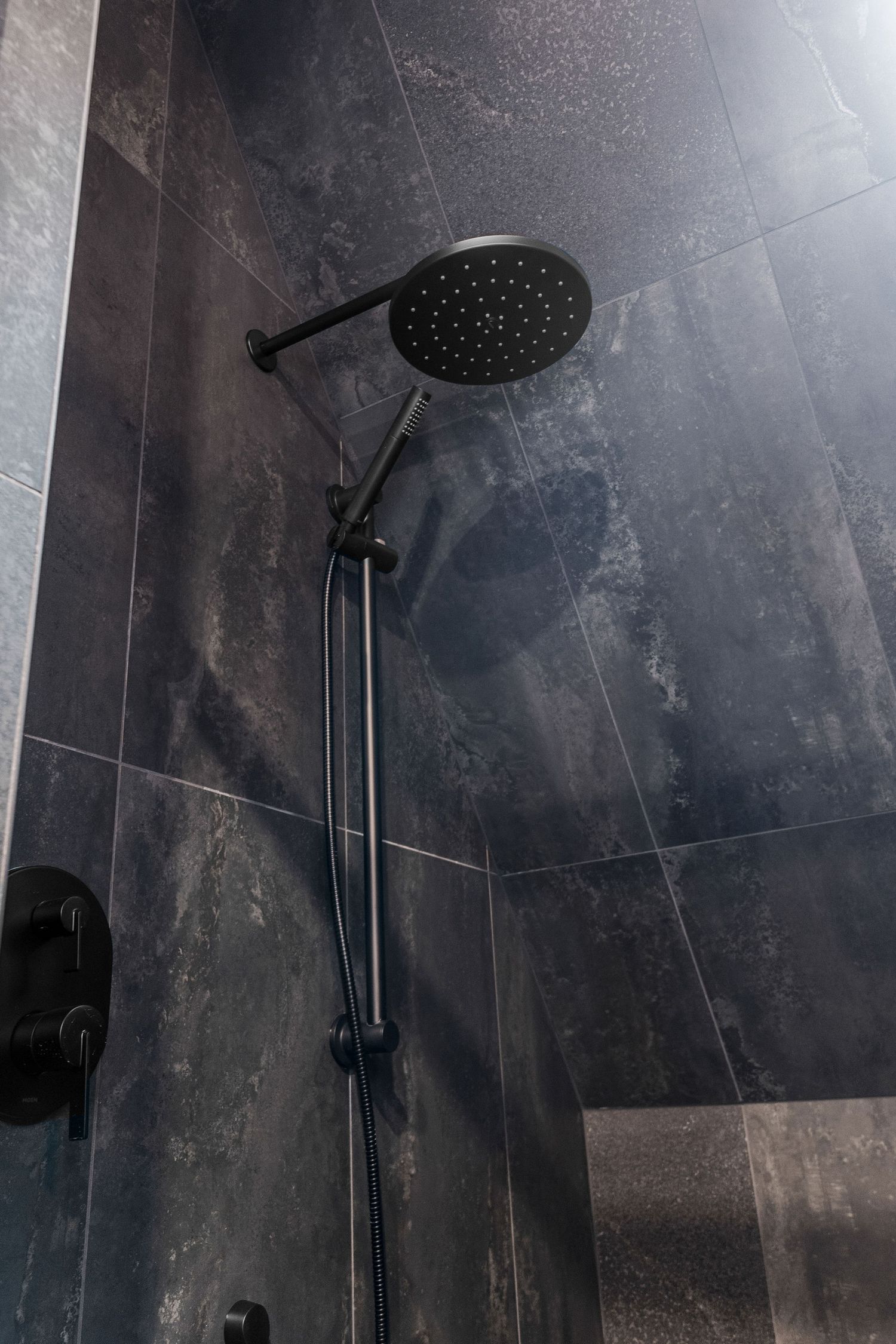 Sleek black rainfall showerhead and handheld sprayer mounted on dark stone tile wall.