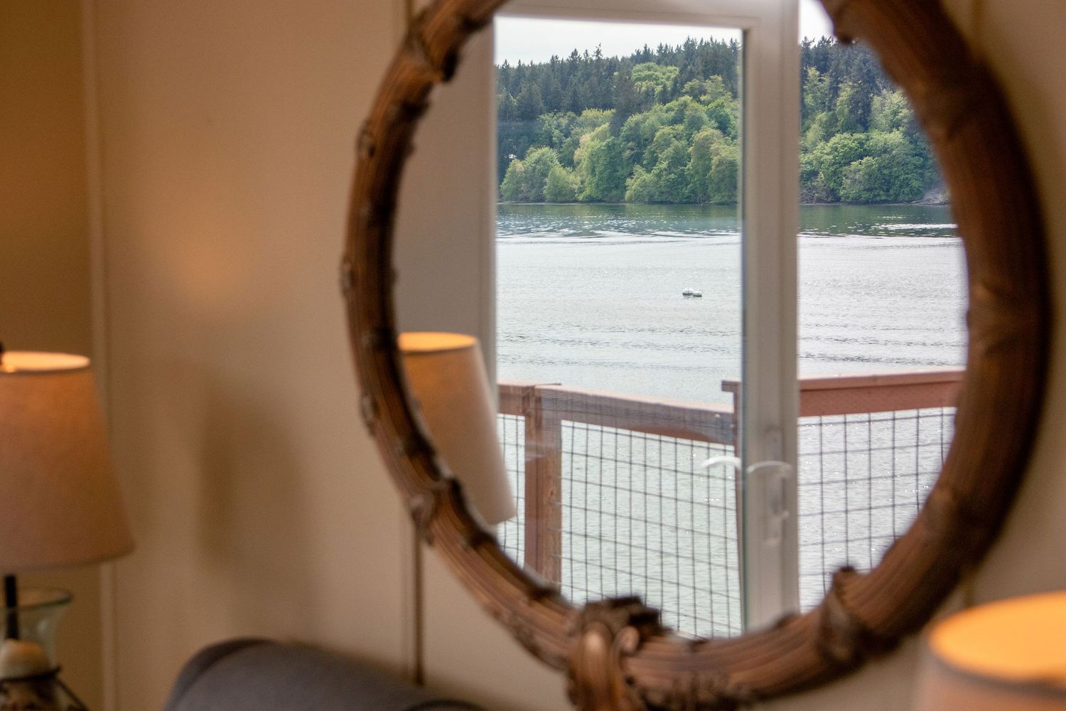 Rustic round mirror on white wall with waterfront view through large windows.