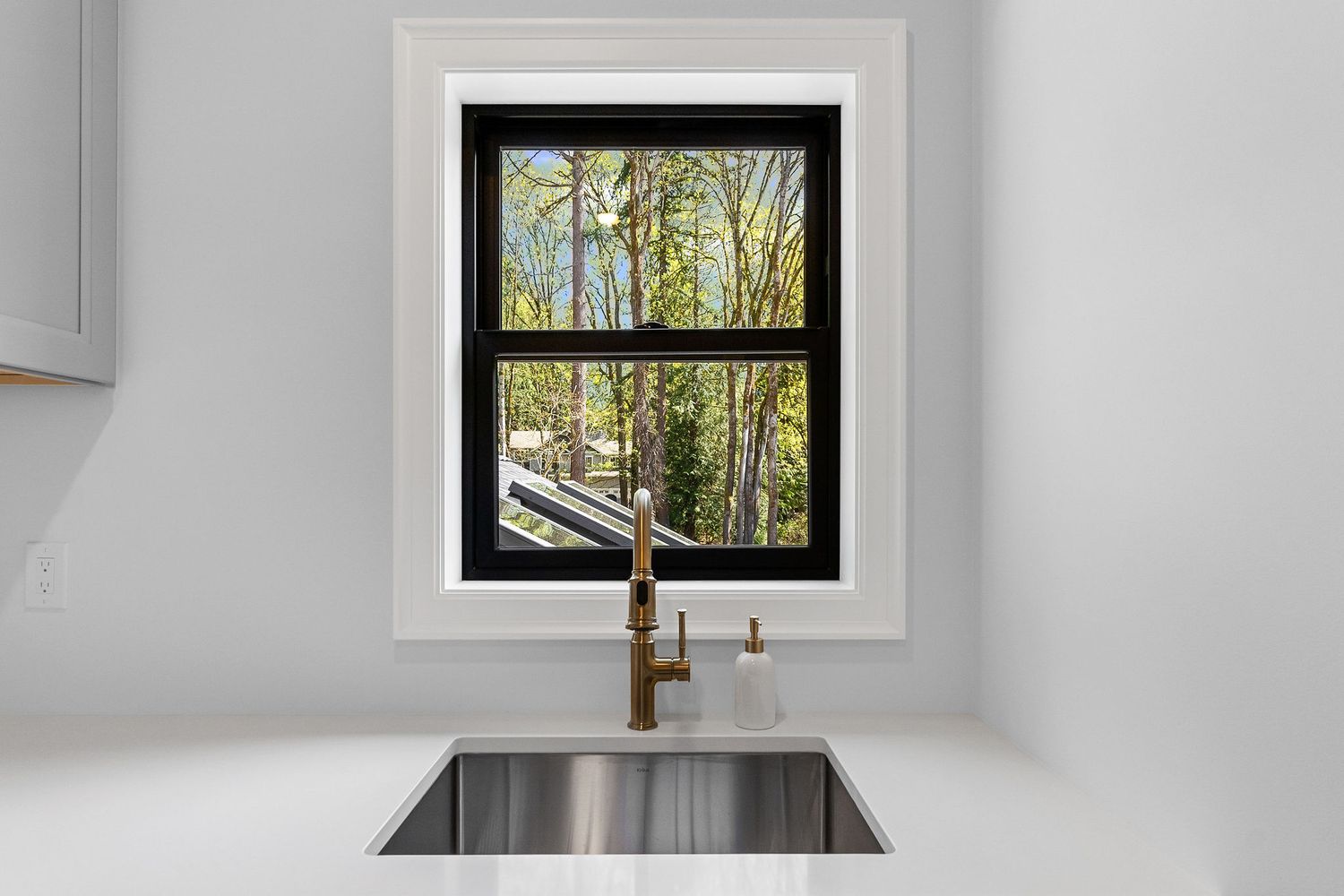 Modern white kitchen with black-framed window overlooking trees and brass faucet over stainless steel sink.