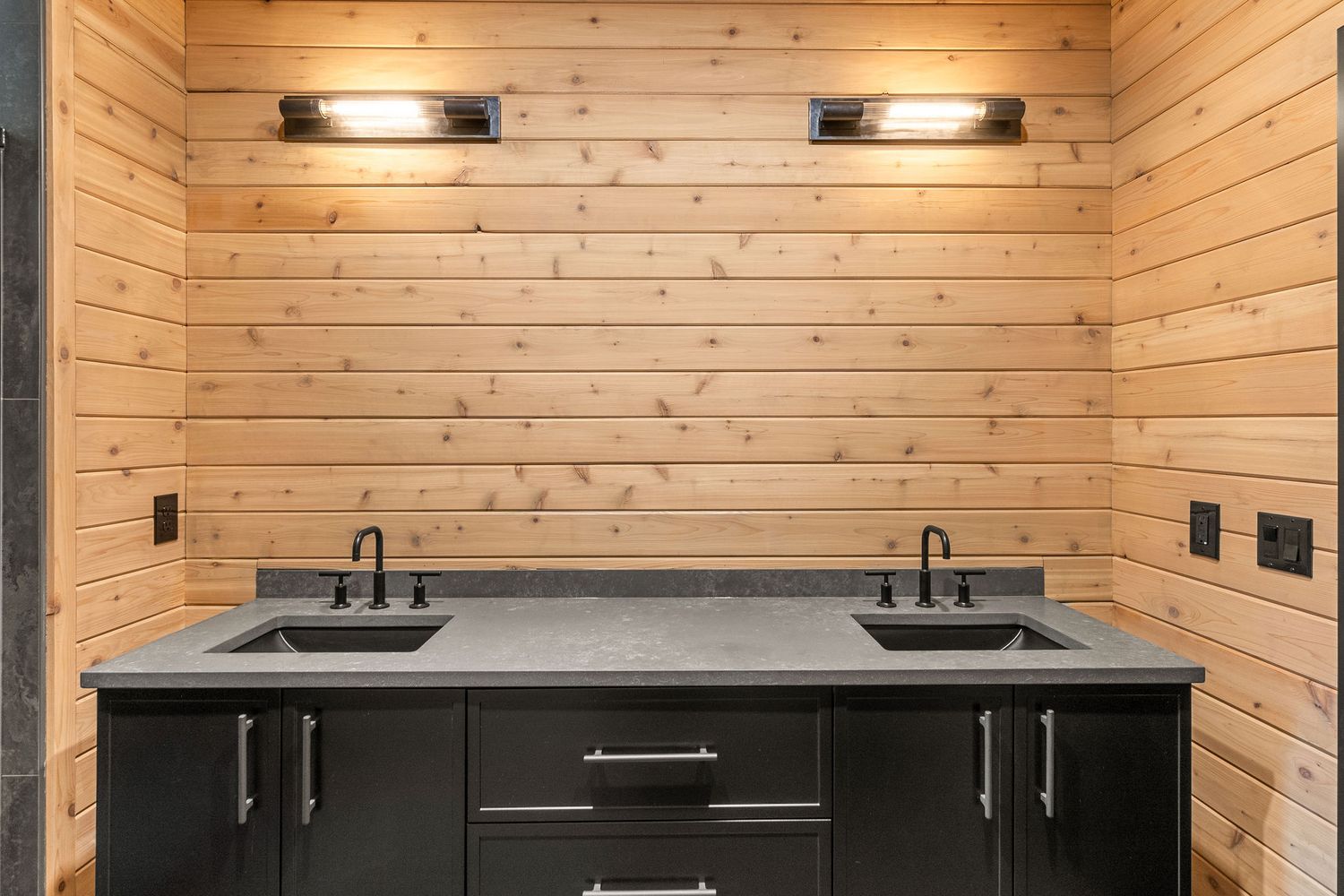 Double sink vanity with black cabinets and grey countertop against pine wood walls.