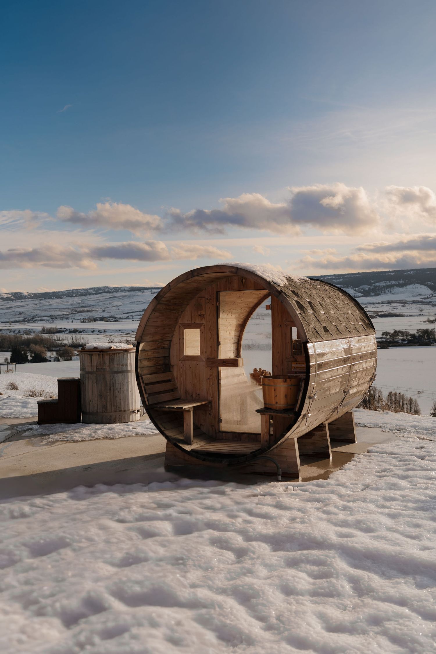 A wooden barrel sauna sits in a snowy landscape with cloudy winter skies and mountains in the background.