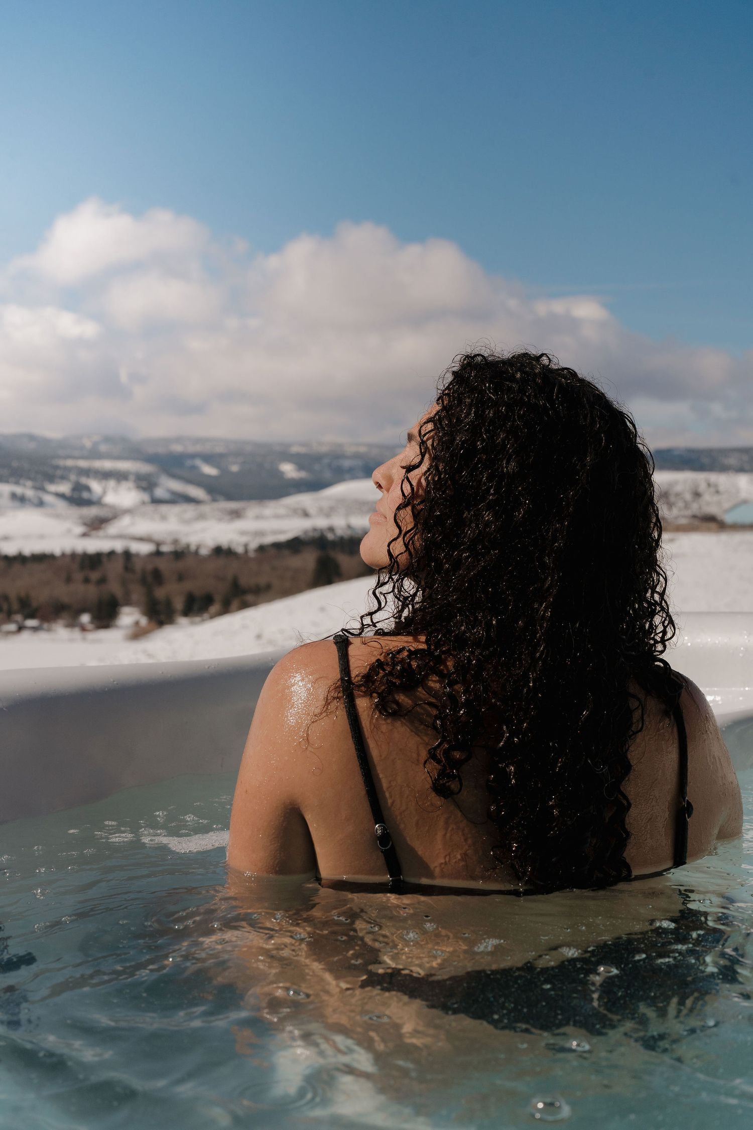 A person soaks in a natural hot spring surrounded by snowy mountains on a bright winter day.