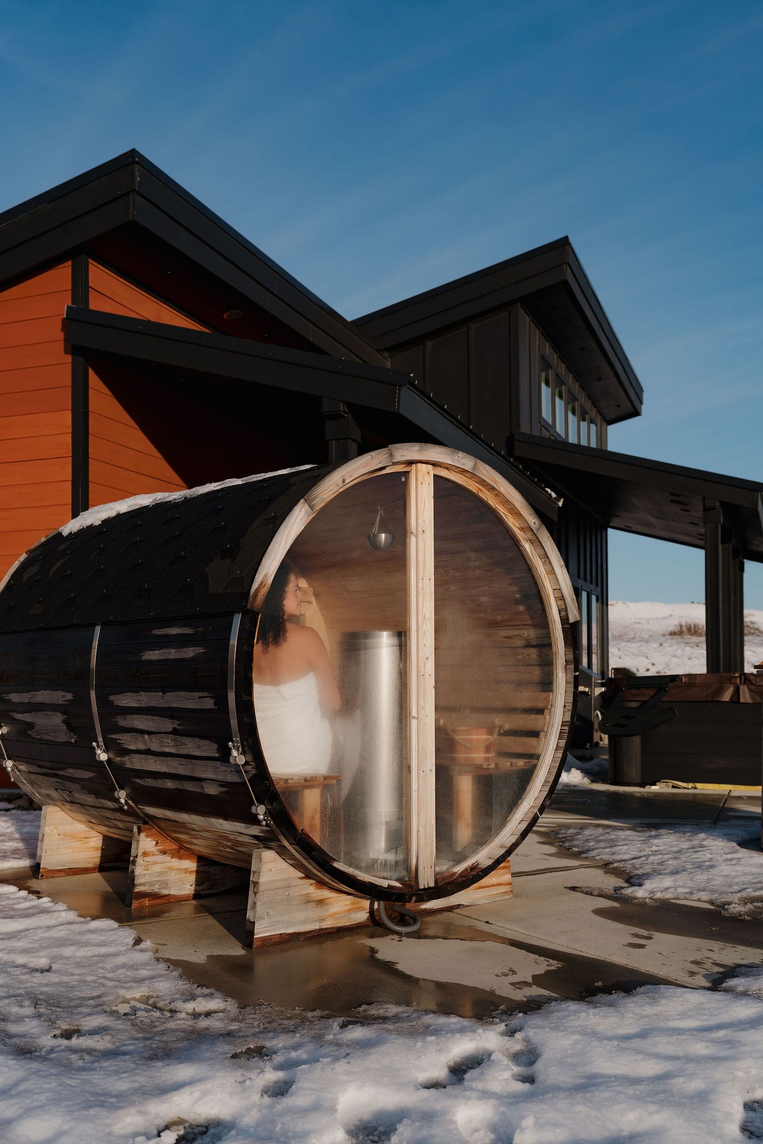Modern wooden cabins with curved architectural elements stand in snowy landscape against blue sky at sunset.
