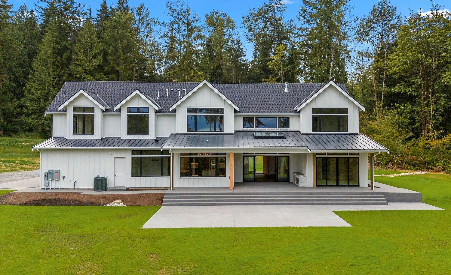 Modern white house with multiple dormers and large concrete patio area in a wooded setting.