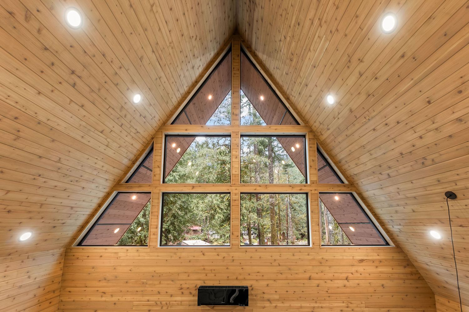 Triangular window wall with forest views in wooden A-frame ceiling with recessed lighting.