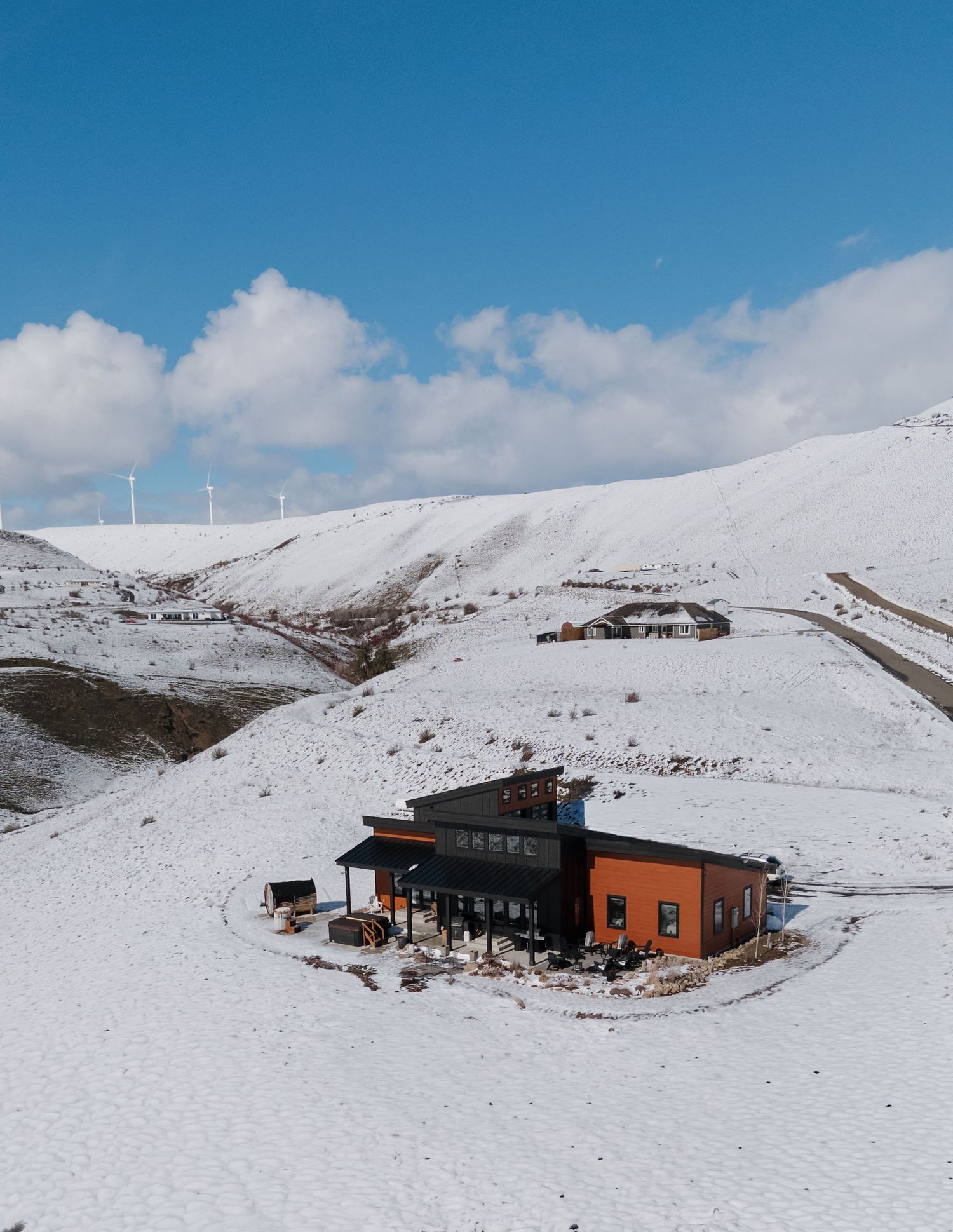 Orange and black building nestled in snowy mountain landscape with wind turbines visible on ridge under blue sky.