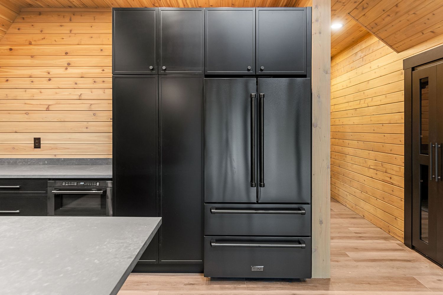 Large black refrigerator with matching cabinets against pine wood walls in modern kitchen.