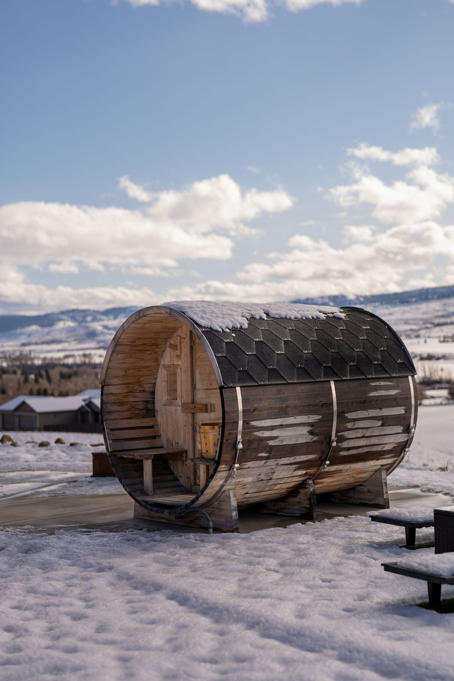 Wooden barrel saunas covered in snow sit along a snowy mountainside with cloudy winter skies above.