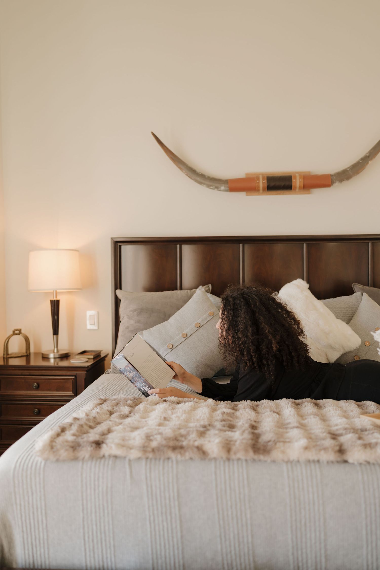Cozy bedroom interior with dark wood headboard, white bedding, decorative bow mounted on wall, and warm lighting.