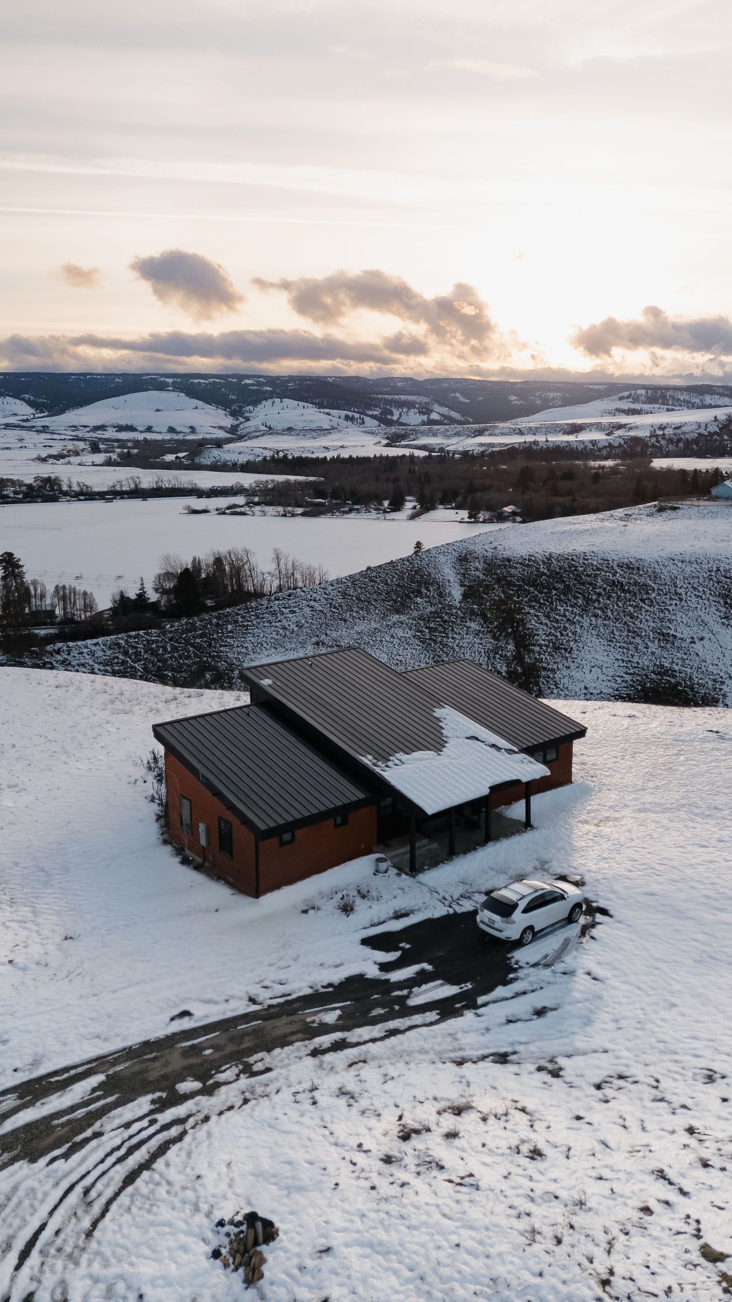 Snow-covered wooden cabins and parked car on a hillside overlooking a winter landscape at sunset.
