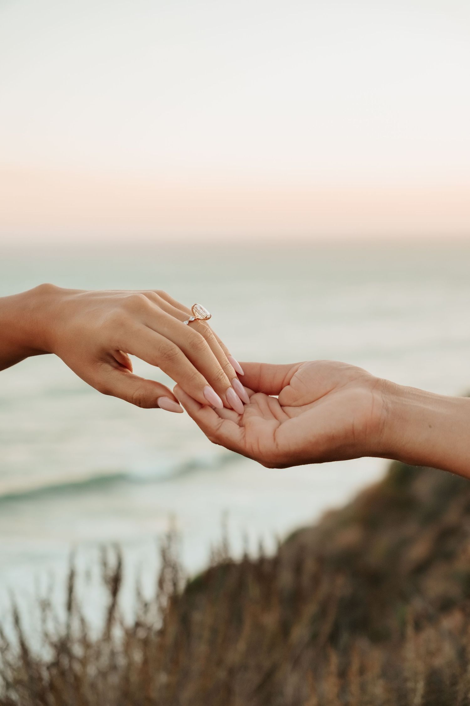 Close-up of couple's hands touching with a beautiful diamond engagement ring near ocean at sunset.