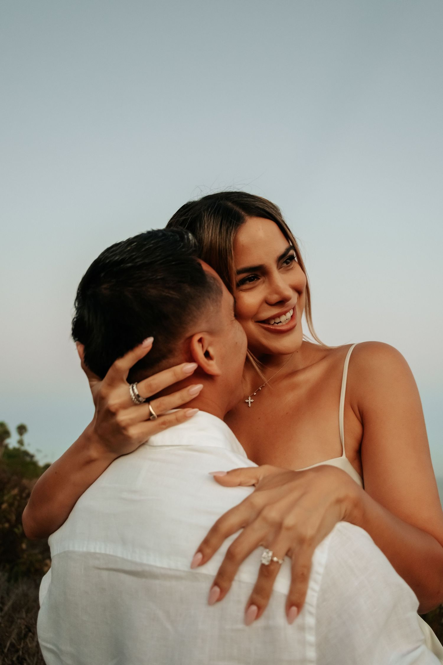 Smiling woman embracing man showing off engagement ring at sunset outdoors.