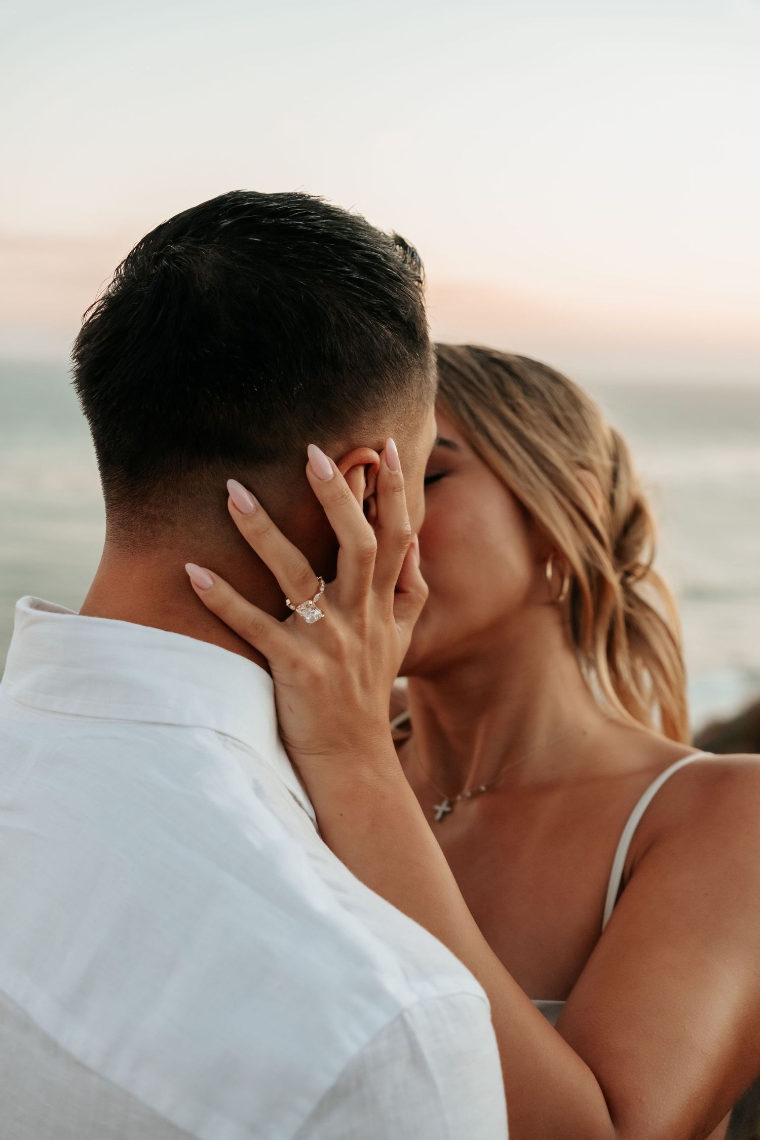 Engaged couple sharing a passionate kiss at sunset beach, woman displaying diamond engagement ring.