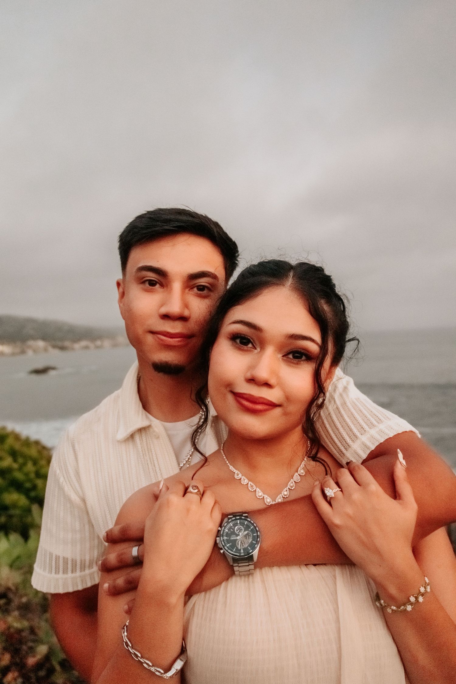 A young couple poses romantically by the ocean, wearing white outfits and silver jewelry under a cloudy sky.